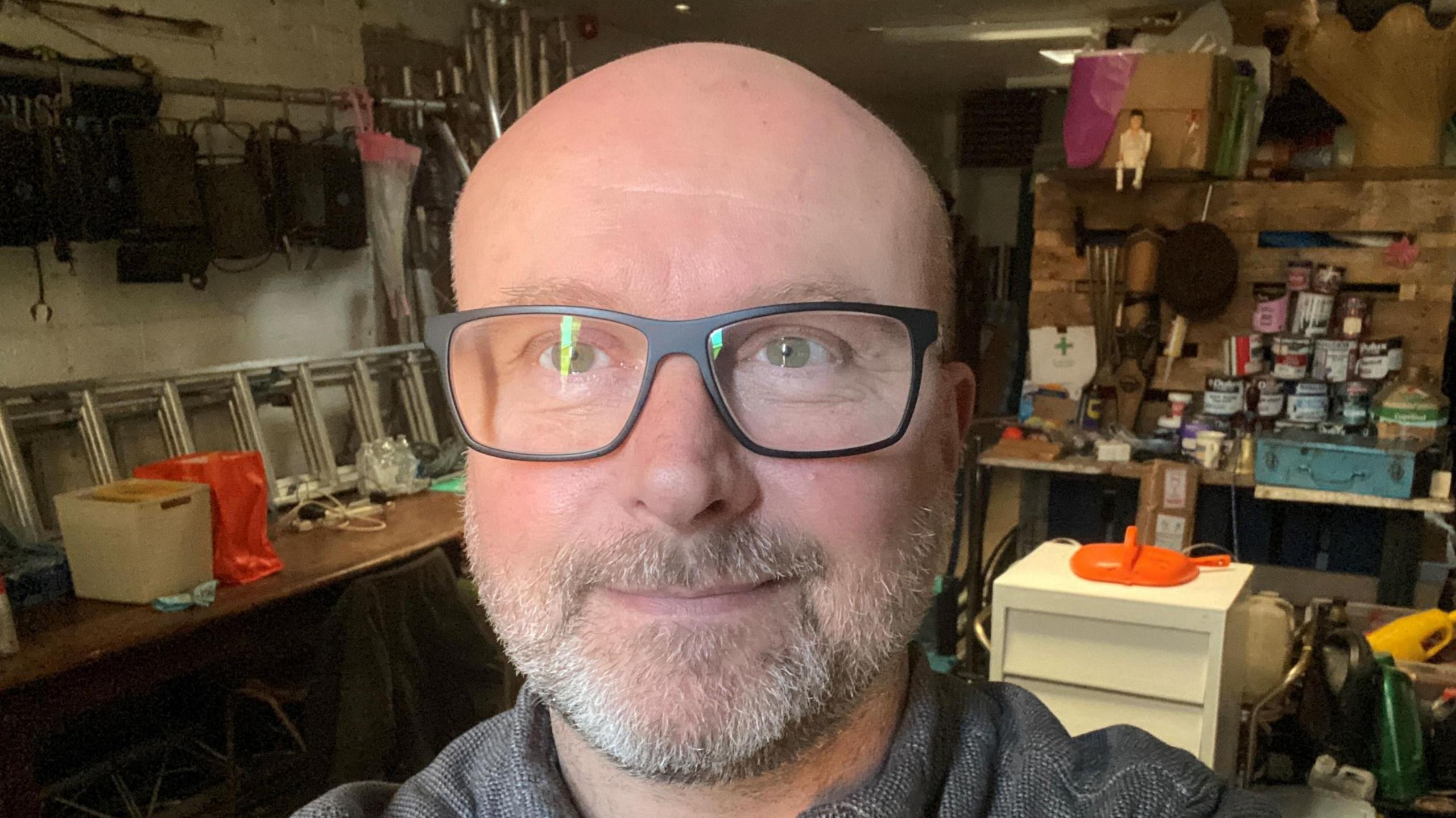 A man with a short grey beard and no head hair standing in a workshop. He has green eyes and black framed glasses on, only his head and shoulders up are visible. He's wearing a dark grey collared top and is smiling at the camera. There are lots of tools, paint cans and DIY equipment in the workshop behind him. 