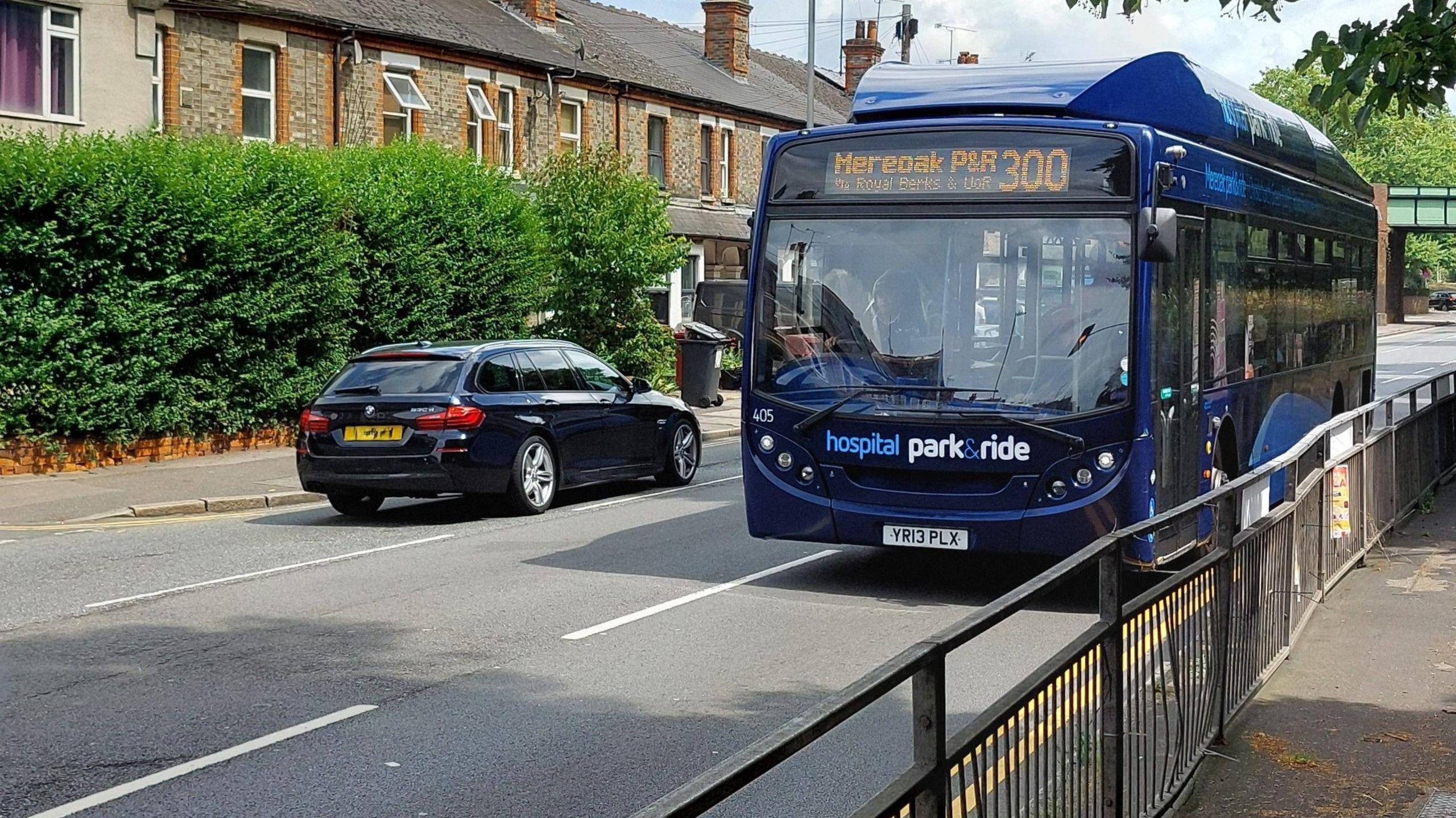 A bus along London Road, Reading, where a new bus lane will be established. It is a blue Hospital Park and Rode bus in a residential area. A dark coloured car is passing by it, travelling in the opposite direction. It is a sunny day.