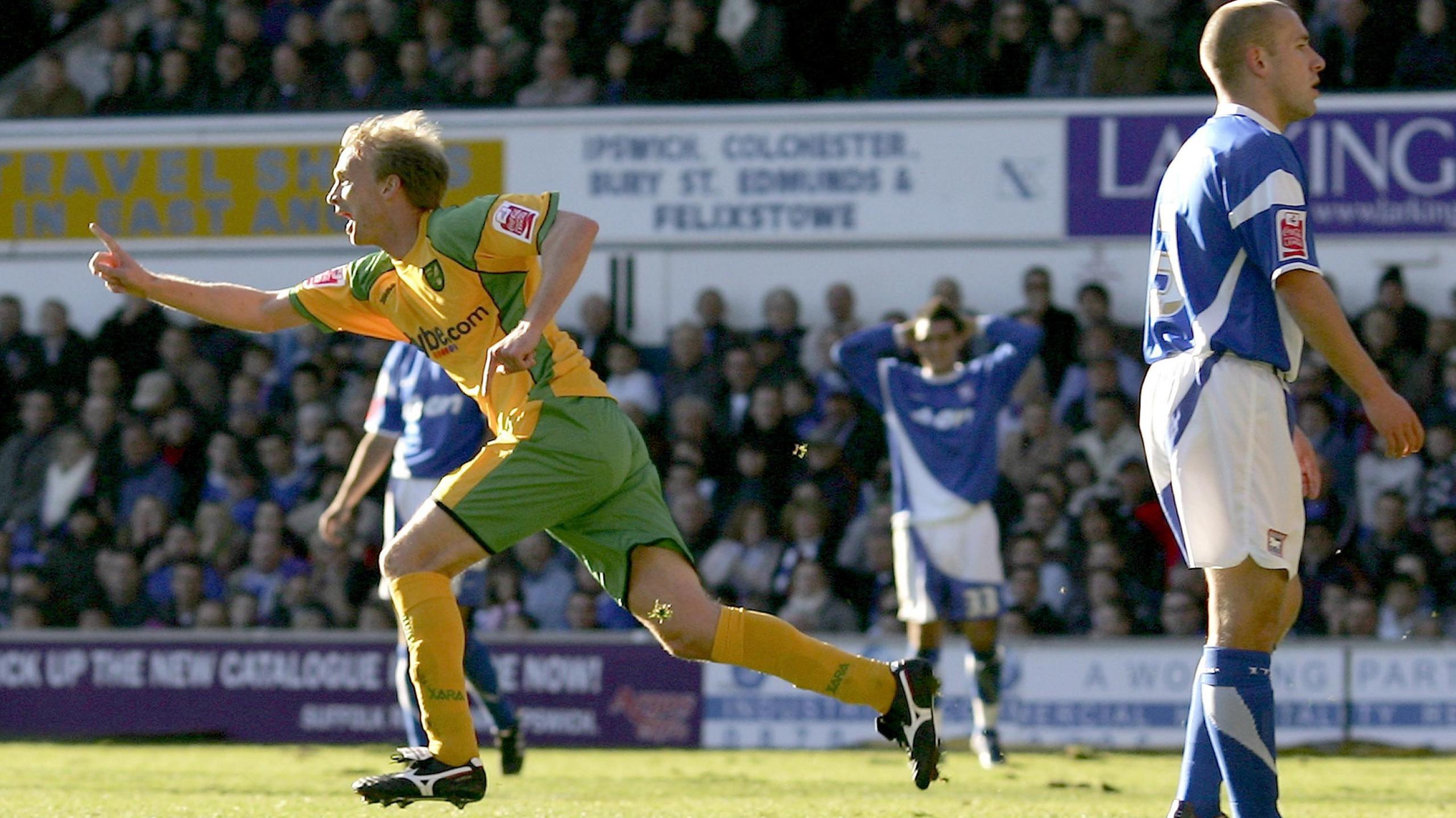 Chadwick, celebrating scoring in the Yellow and Green of Norwich City. He has one finger raised in celebration, as an Ipswich Town player, in full blue and white kit, looks dejected at conceding at Portman Road.
