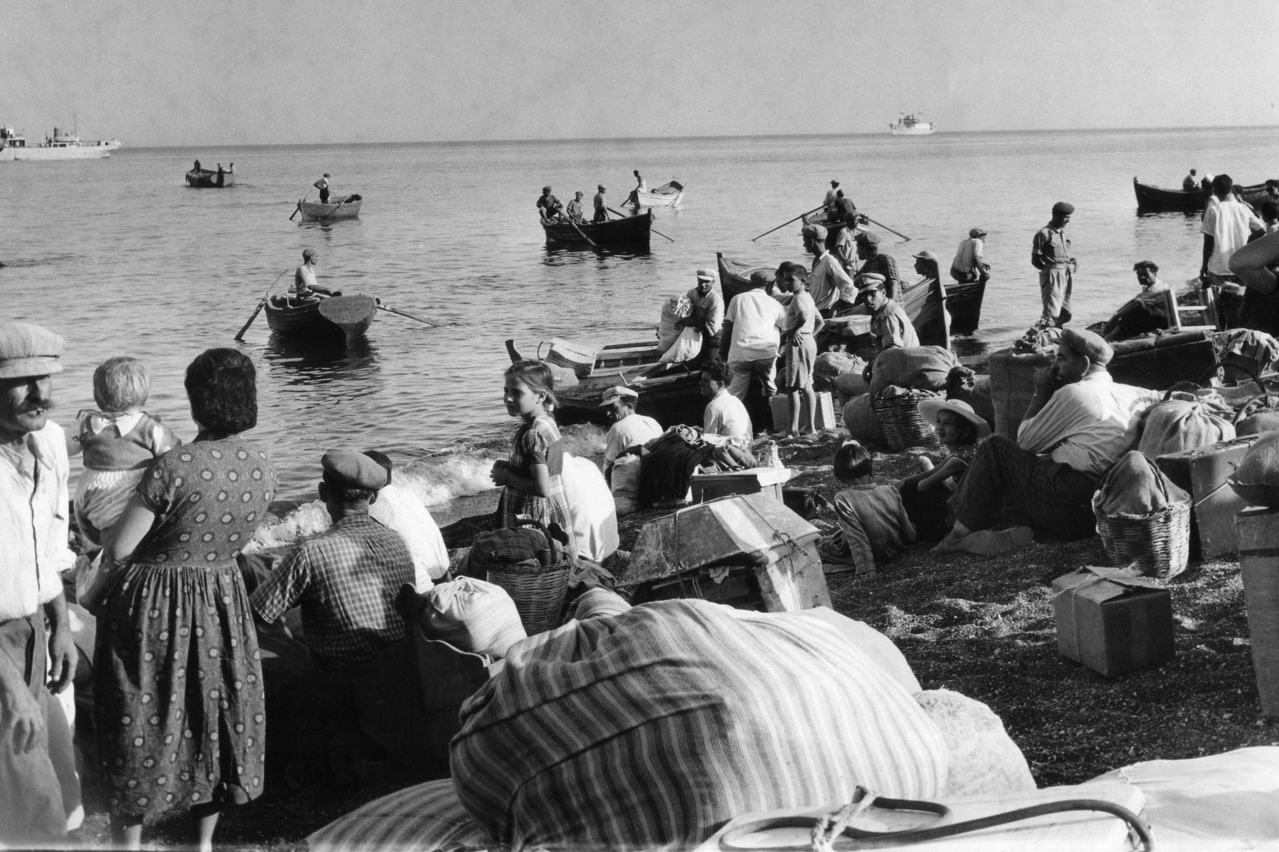 A black and white photo showing families with bags gathered on a beach. Some of them are rowing into the distances in small wooden row boats