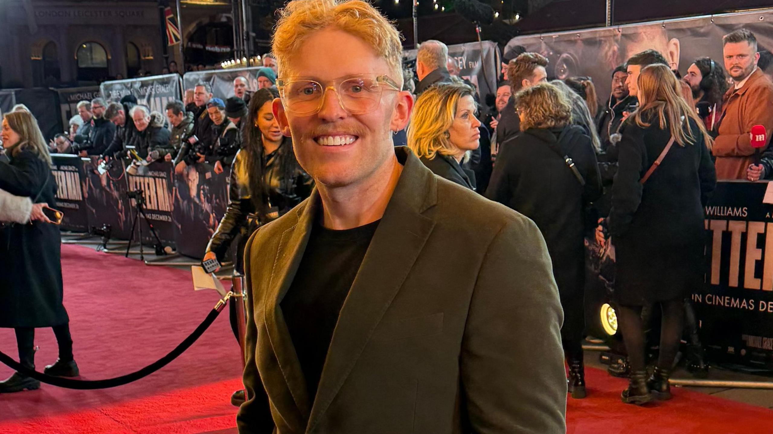 Adam Tucker wearing a suit blazer and black t-shirt stood on the London red carpet premiere of Better Man. Fans and press are stood on the other side of a barrier behind him.