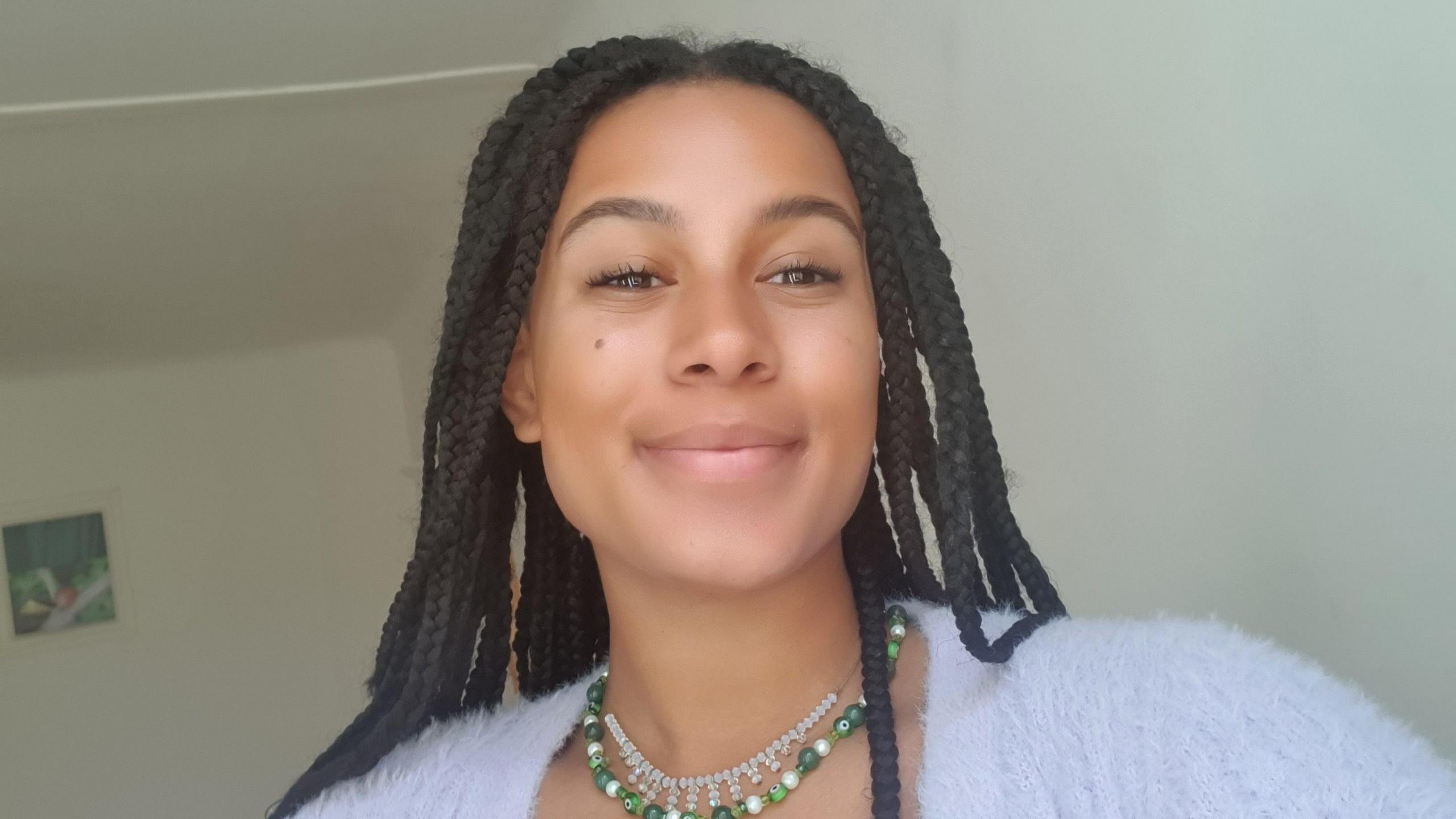 Darwin Arnold smiles at the camera while taking a selfie inside a room painted white. She wears a fluffy pale purple cardigan and a green beaded necklace, her braided hair worn loose over her shoulders. 