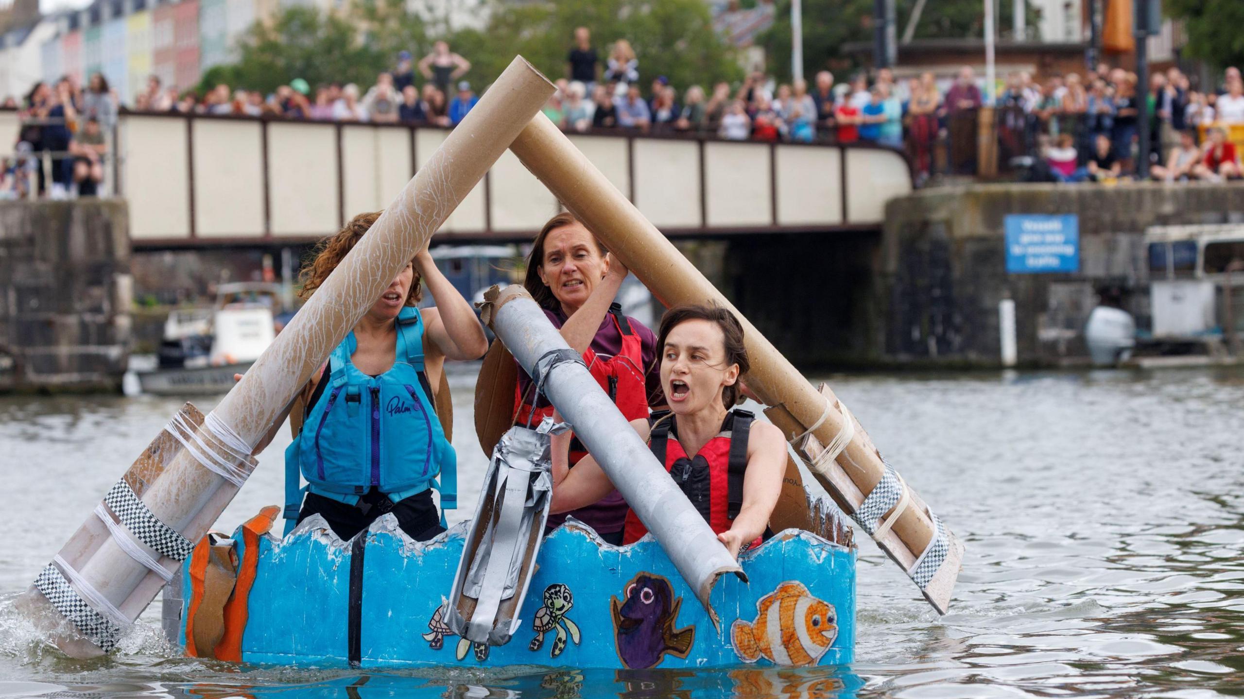 A trio of young women paddle their homemade cardboard craft on Bristol's harbourside during the Bristol Harbour Festival 2024. In the background large crowds can be seen watching from the Prince Street Bridge. The women are using large cardboard oars and their craft is painted blue and decorated with film characters such as Nemo and Dory.