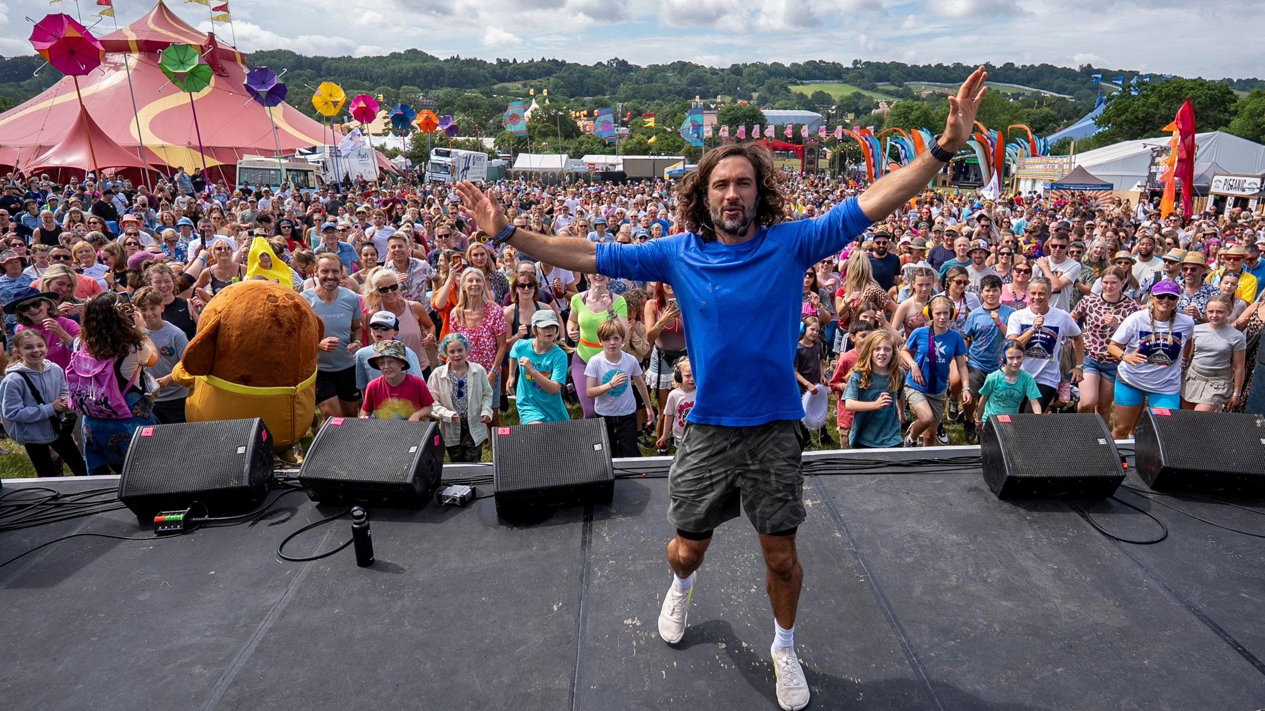 A man wearing a blue long t-shirt in front of a huge crowd of people while standing stage