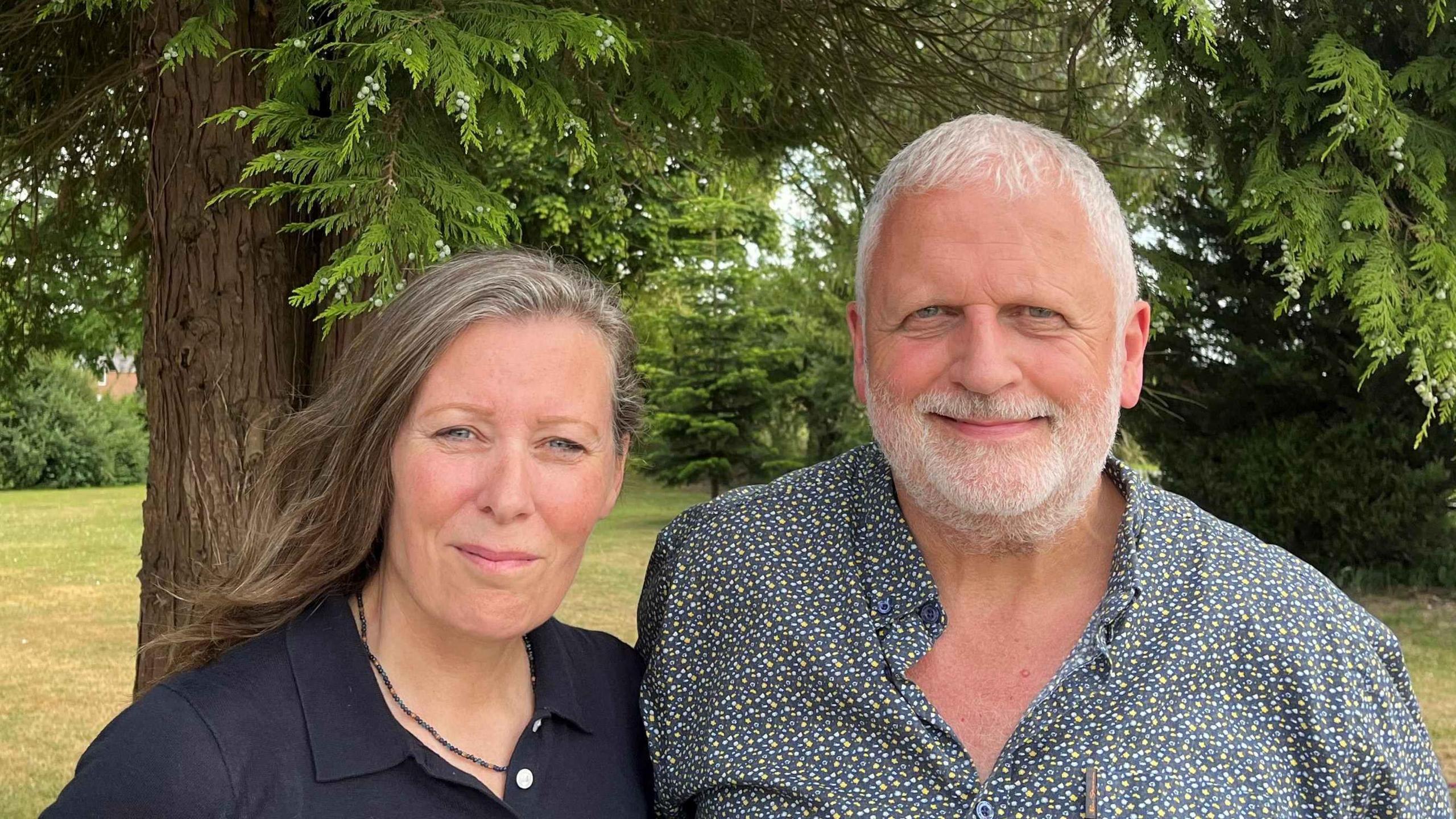 Moya Hampson and Mike Daniel standing under a tree. Moya is wearing a black shirt and has long dark hair. Mike is wearing a patterned shirt and has white hair.