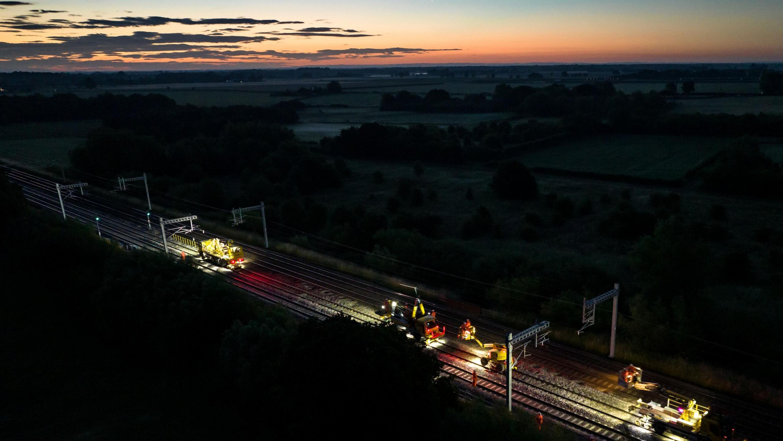 Work taking place overnight on rail lines. Machines can be seen on the track. In the background the sun is setting over the horizon