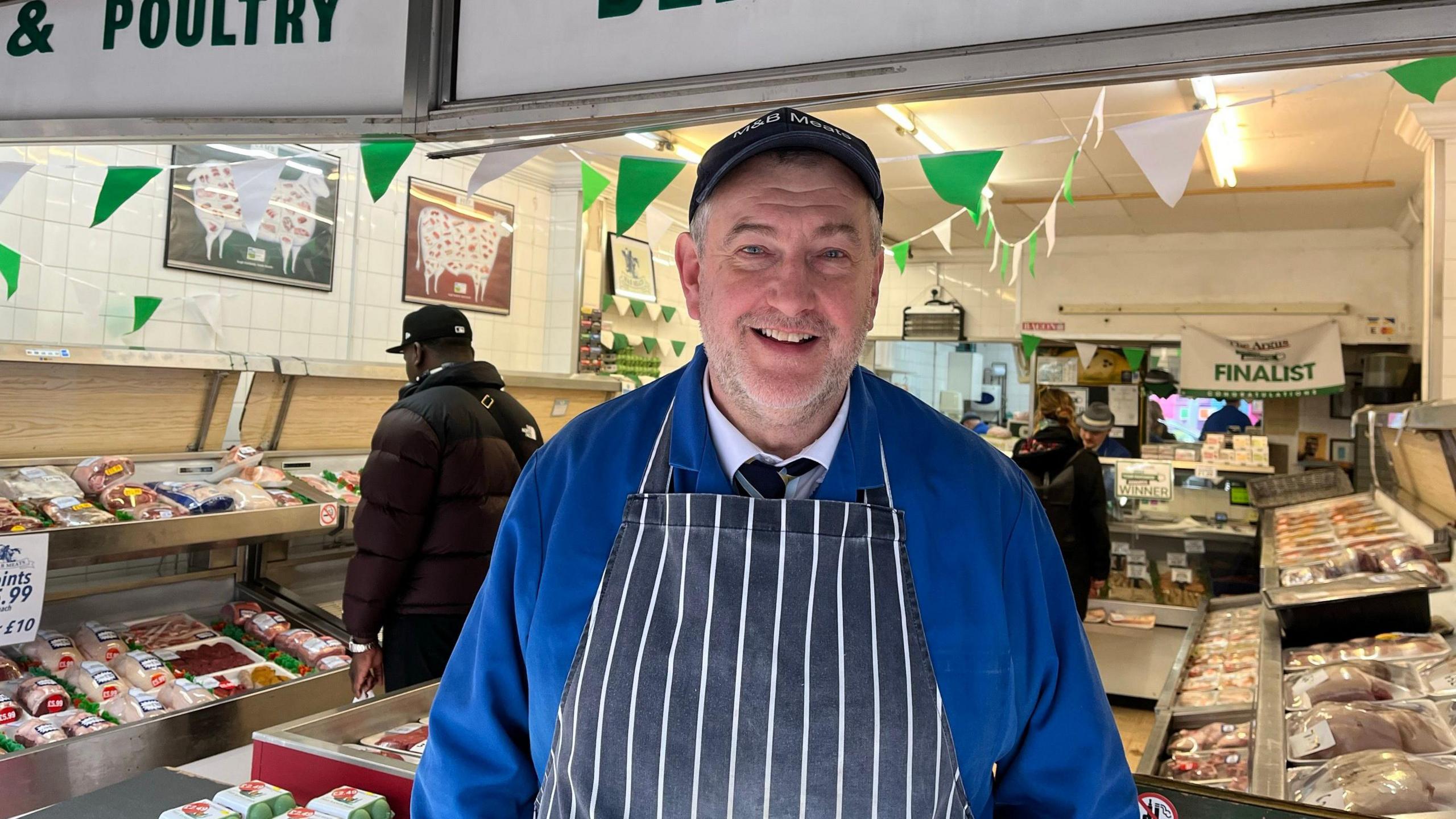 A middle aged man wearing a cap and royal blue butchers coat and blue and white striped apron smiles at the camera in front of his butchers shop in Brighton