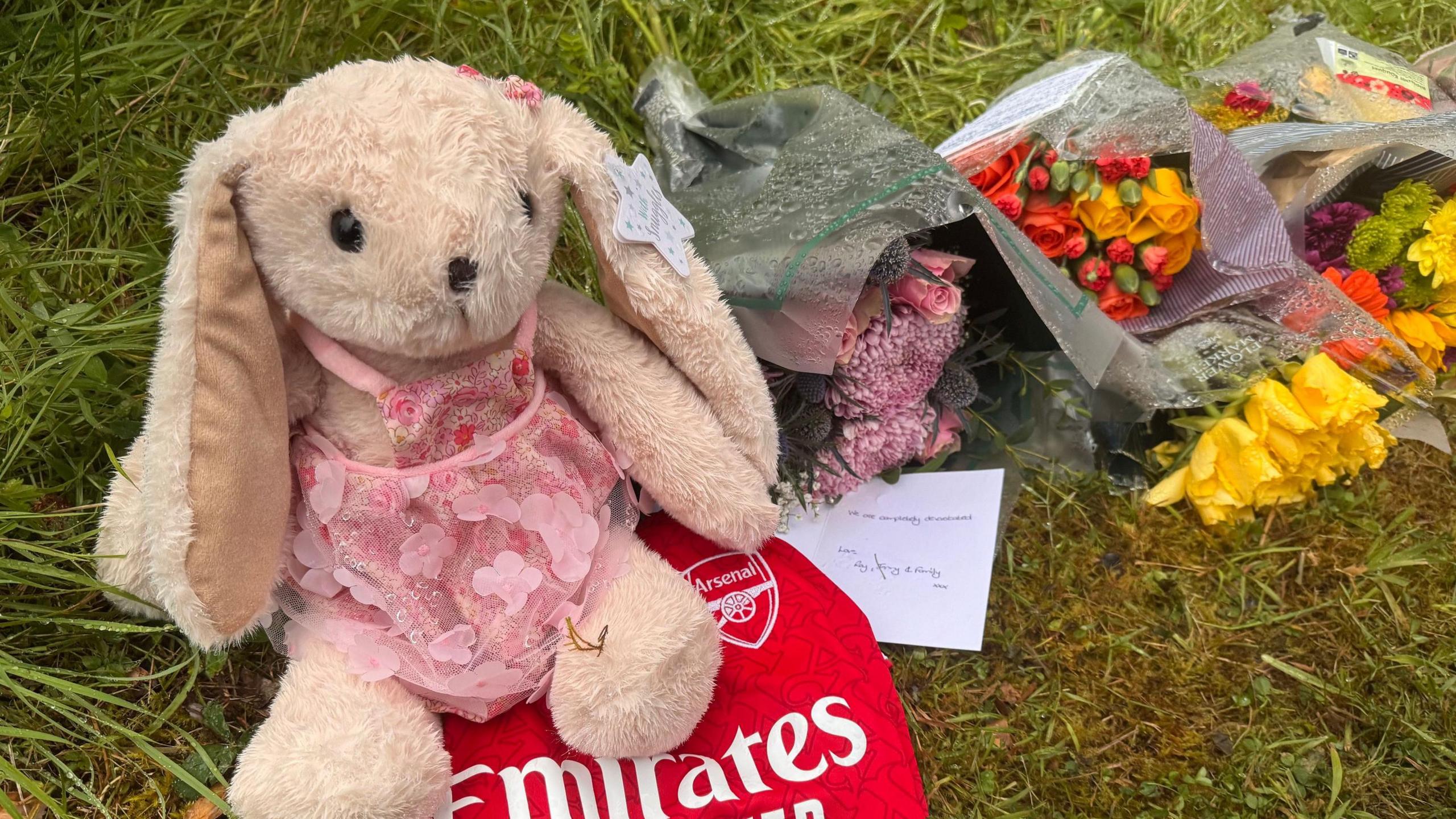 A collection of gifts and flowers left near the scene of the fatal shooting in Maguiresbridge. A stuffed toy rabbit wearing a pink dress is sitting on a red Arsenal football top on a patch of grass. Bunches of flowers are lying nearby.
