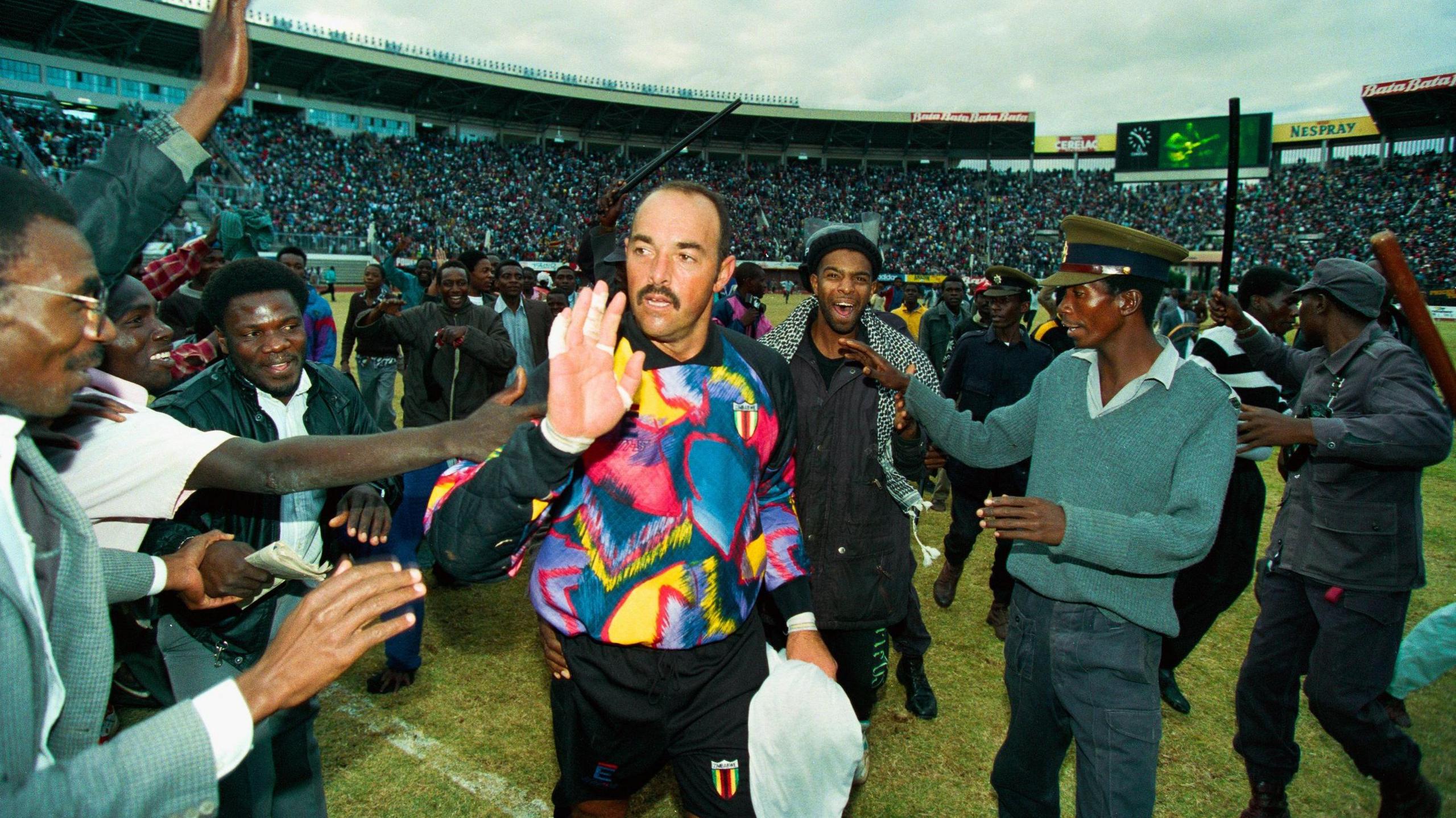 Goalkeeper Bruce Grobbelaar wears a bright, multicoloured jersey as he walks off the pitch and raises his hand to acknowledge the applause of fans and security staff after a World Cup qualifying match between Zimbabwe and Cameroon in 1993 in Harare.