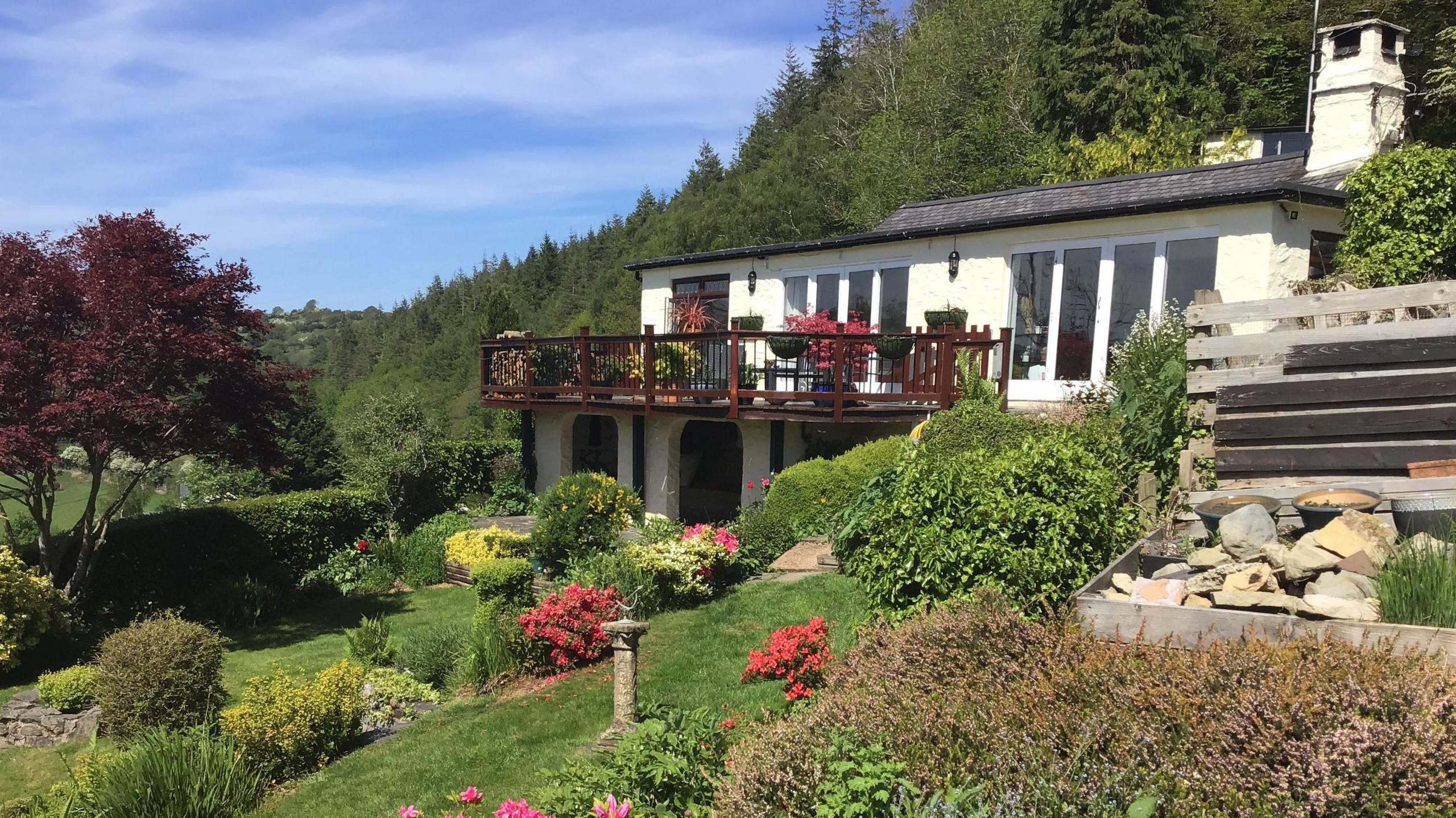 A view of the house, with a tiered garden. It is a white building on one level with a mahogany coloured balcony and a forest behind it