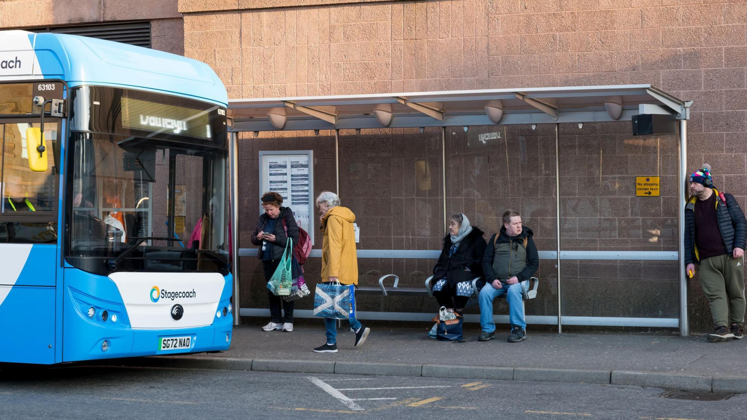 A blue and white Stagecoach bus pulls up at a bus stop in Inverness city centre. A woman wearing a yellow coat walks past the bus shelter where a woman is standing and a man and woman are sitting on a bench. A man wearing a woolly hat with a pom-pom leans against the bus shelter.