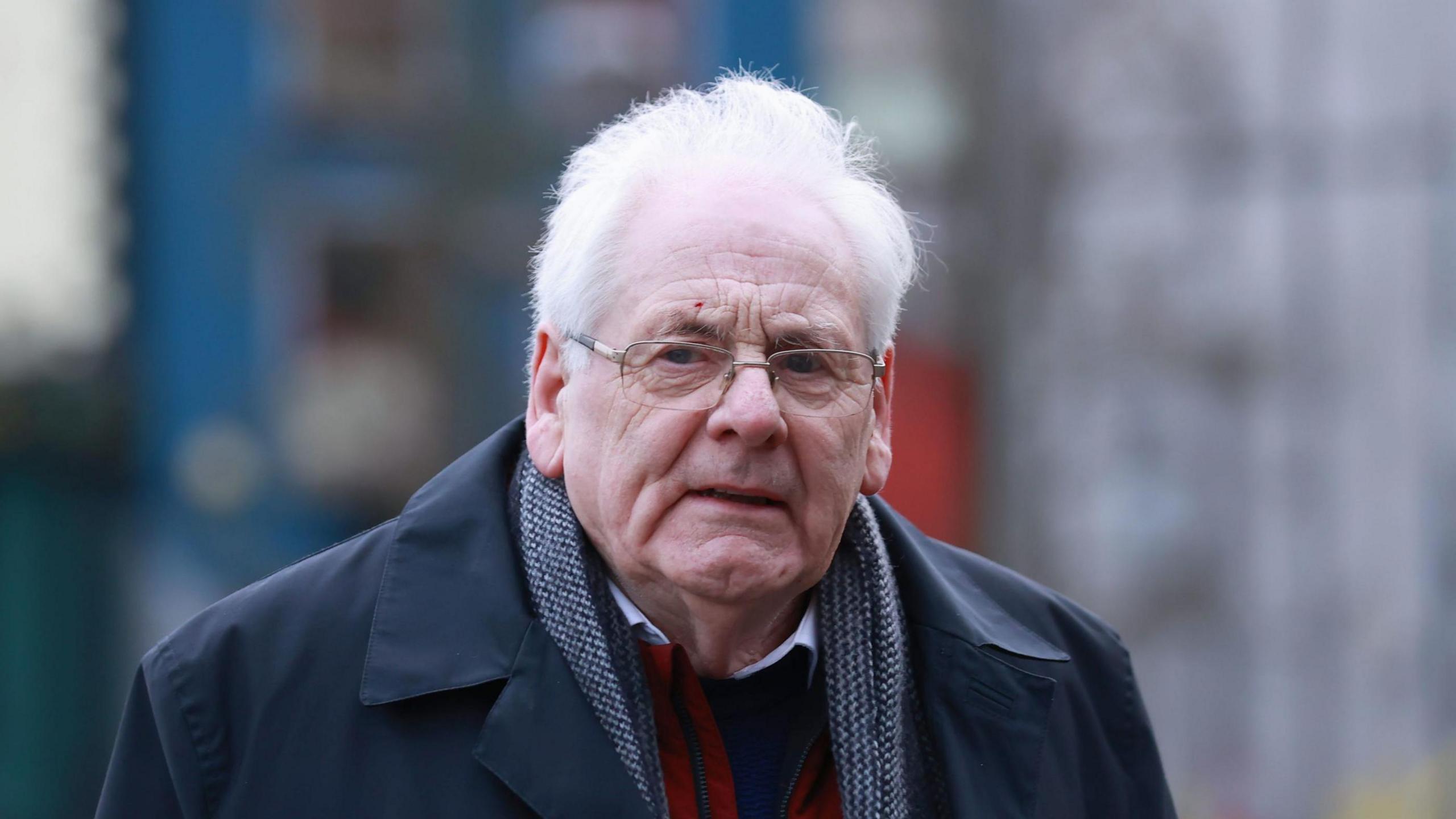 Michael Gallagher has grey hair and is wearing metal-framed glasses. He is wearing a navy overcoat, grey scarf, red gilet and a shirt and jumper.