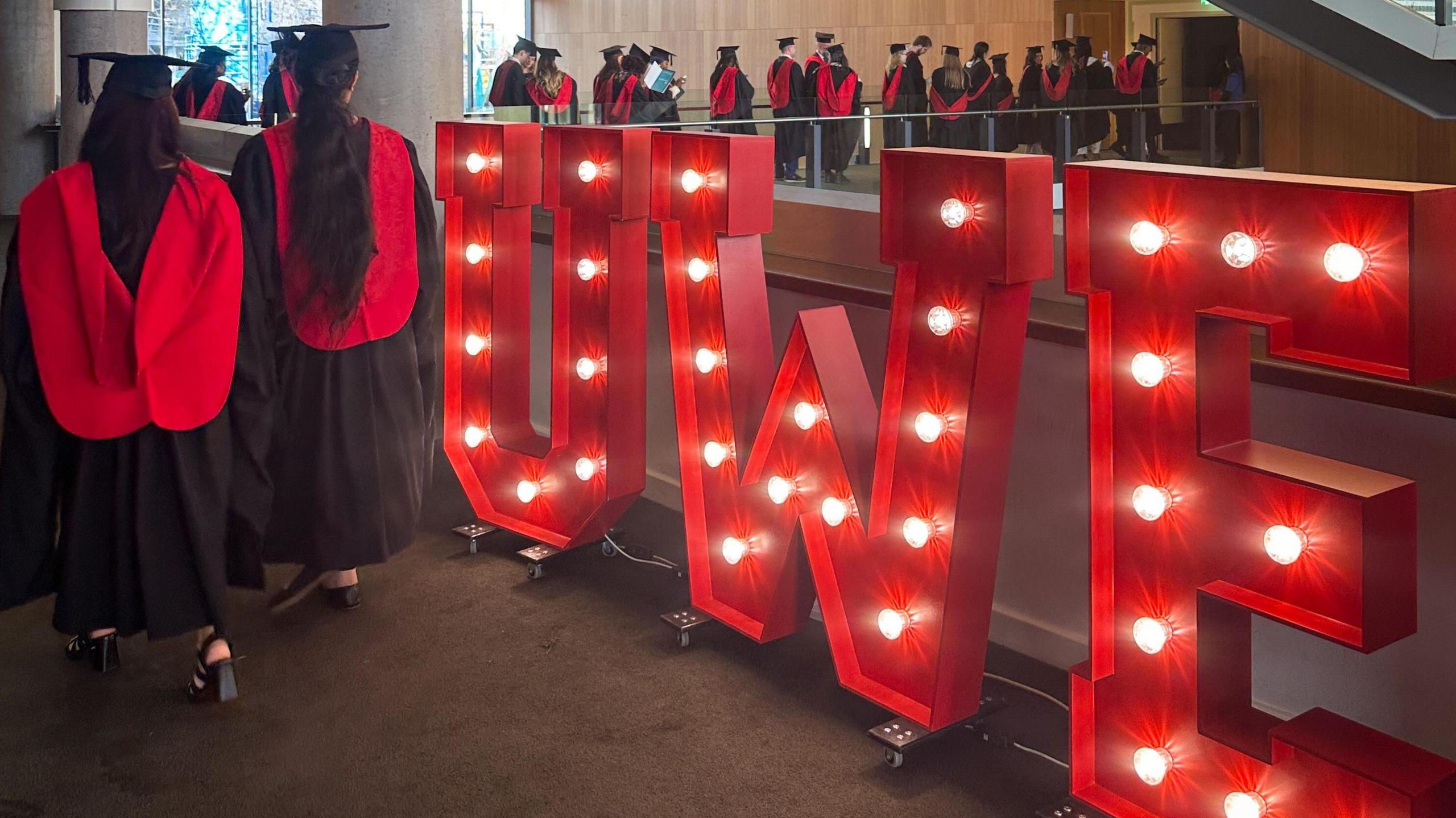 A group of graduating students with black and red gowns walk past a large red illuminated series of letters spelling out "UWE". The students are queuing along one of the floors of the Bristol Beacon to enter the main auditorium