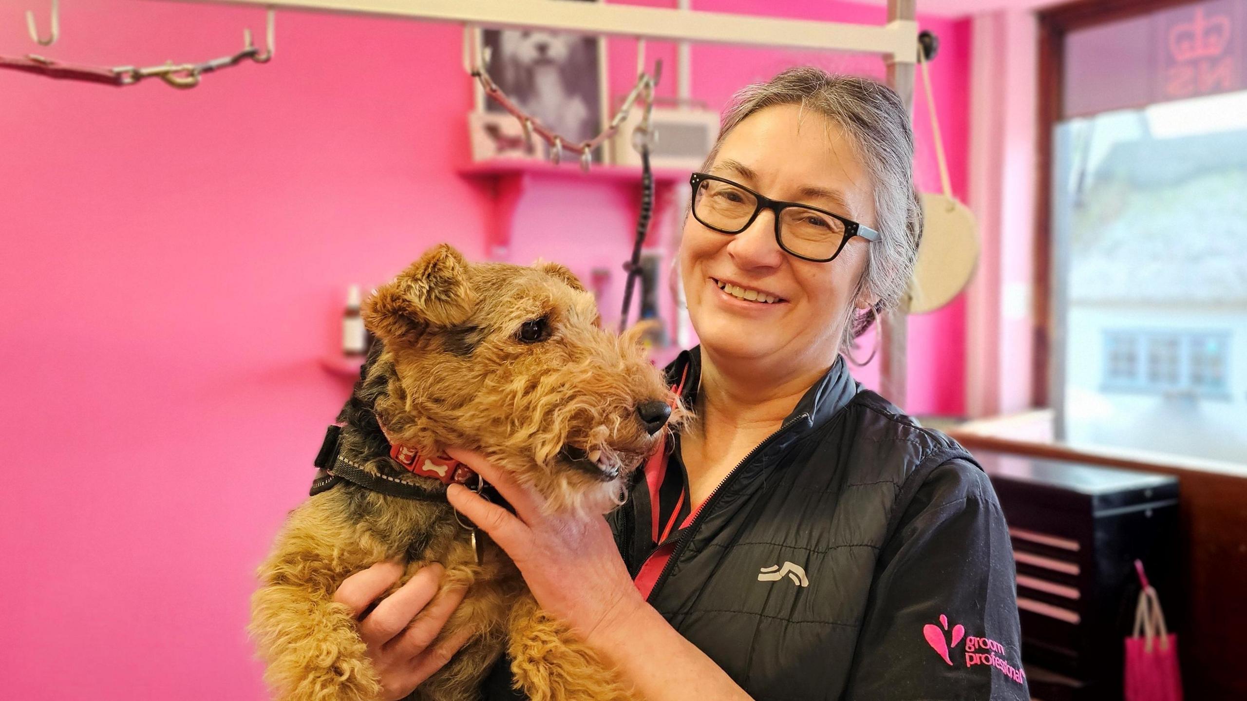 Silvi Donges is smiling at the camera while holding a Welsh terrier. She is standing in her grooming parlour, which is very pink.