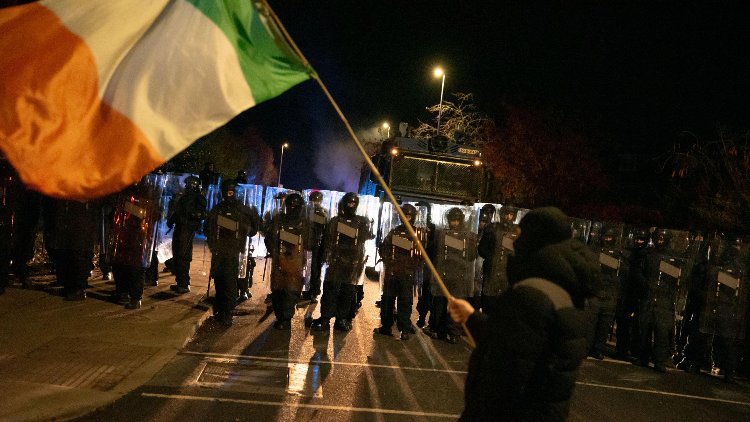 A person clothes in black is flying an Irish flag on a stick. In front of him s a large group of armed police officers, they all have shields and are wearing helmets. The sky is dark. There is also a police truck in the back ground. 