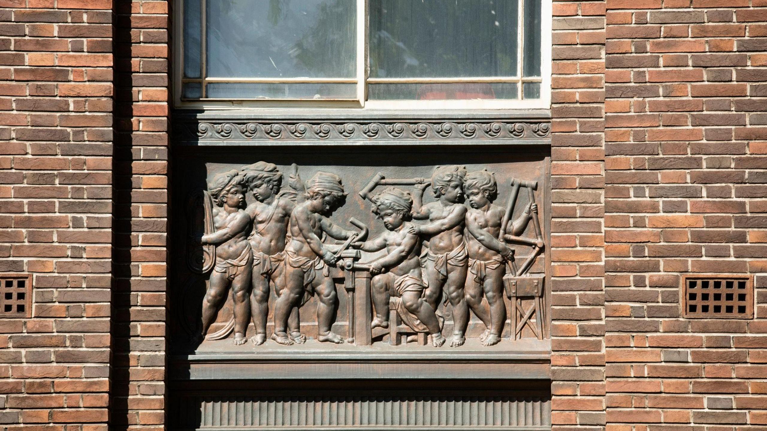 A bronze  plaque set into the wall of the building, showing six child figure working on parts of a bicycle