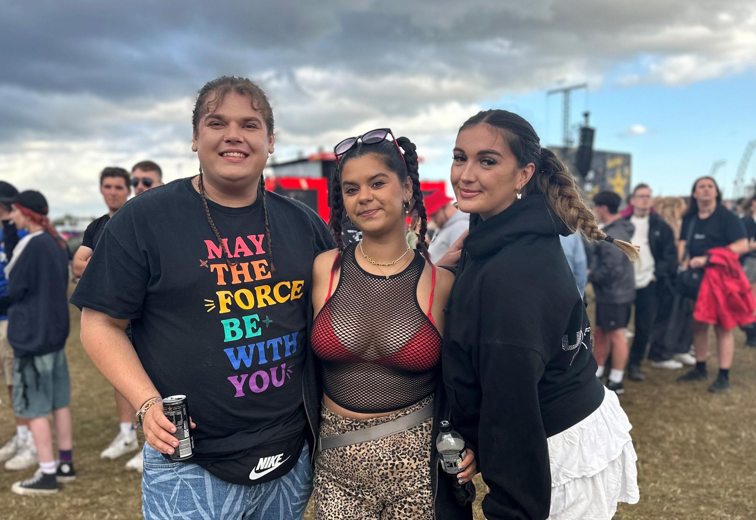 A man and two women stand in a field in front of the main stage at Reading Festival. They're holding drinks and smiling.