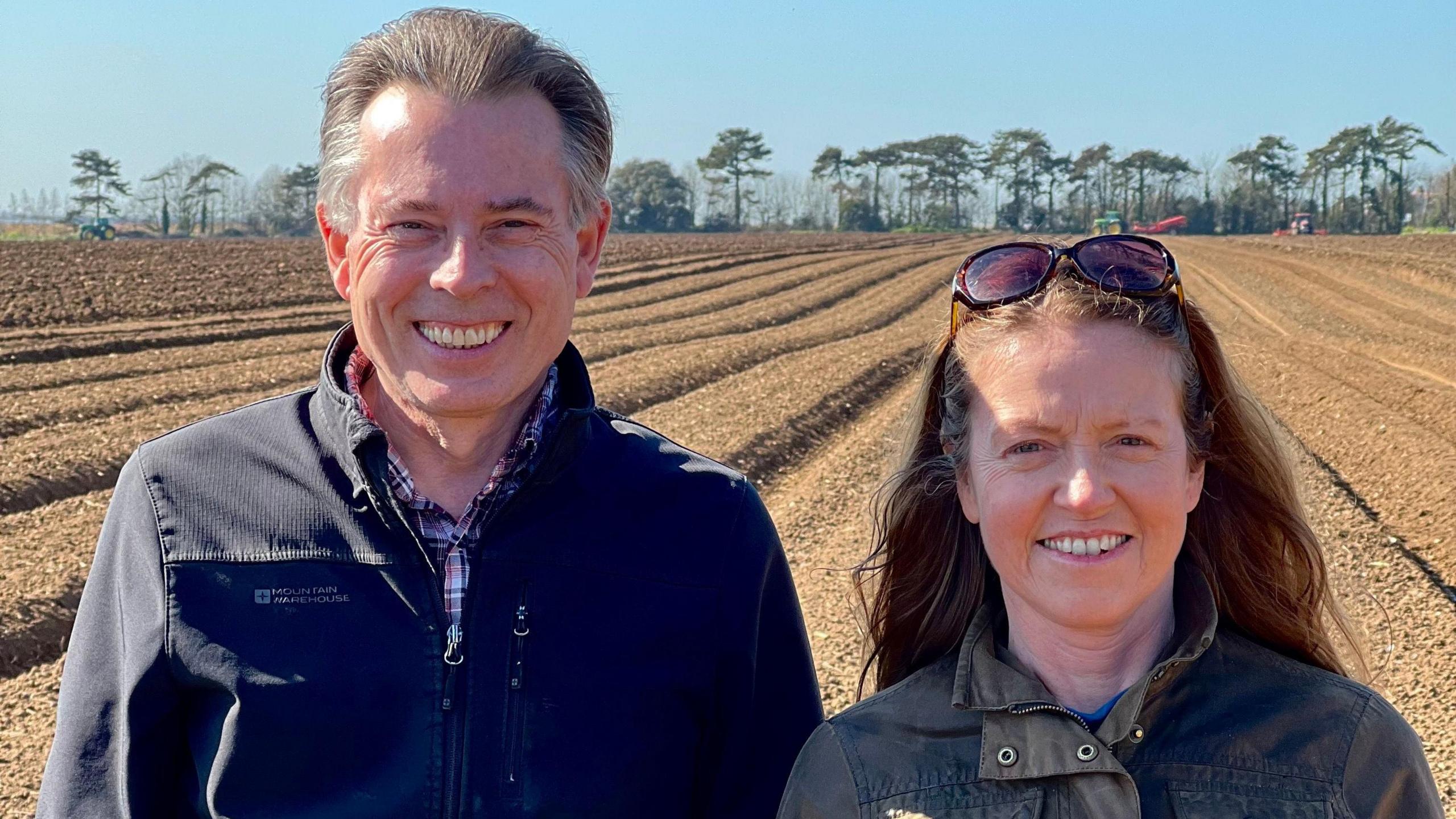 A middle-aged man and woman are stood next to each other in the foreground of the picture. The man has short grey hair and is wearing a jacket over a check shirt. To his left is the woman, with sunglasses on her head and long brown hair, also wearing a jacket. Both are smiling and looking at the camera. In the background is a ploughed field, with agricultural machinery and a line of trees visible in the distance. The sky is clear and blue.