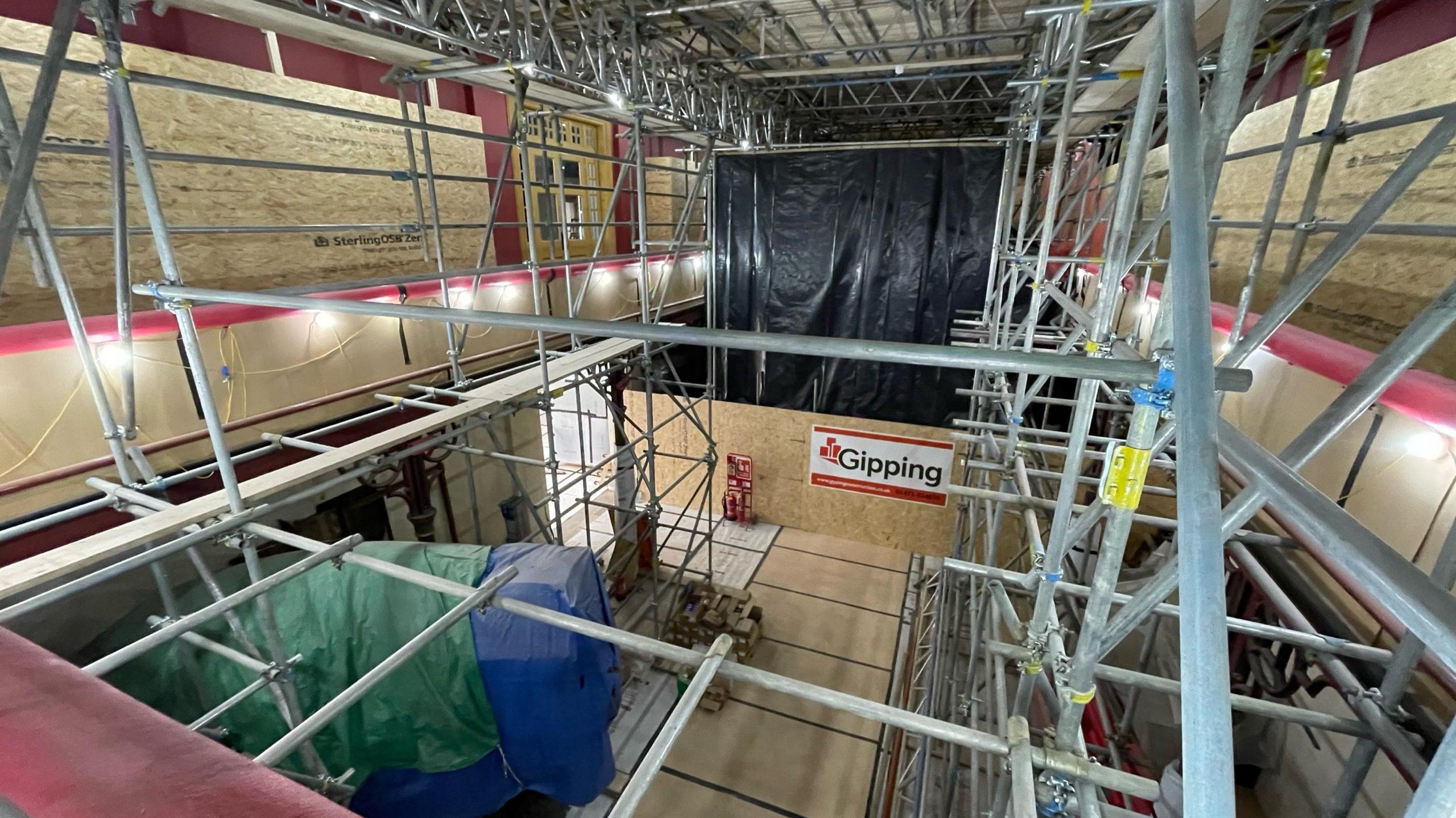 A high view of the Central Victorian Natural History Gallery from a balcony. The covered mammoth can be seen below while scaffolding surrounds the entirety of the room. In the distance a black plastic sheet covers the wall.