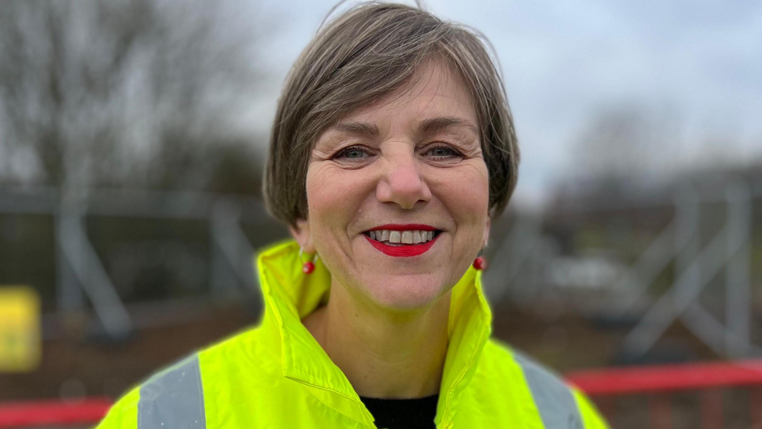 Lilian Greenwood is a woman with short hair is looking at the camera and smiling. She is wearing a yellow fluorescent jacket and has red earrings and red lipstick on. 