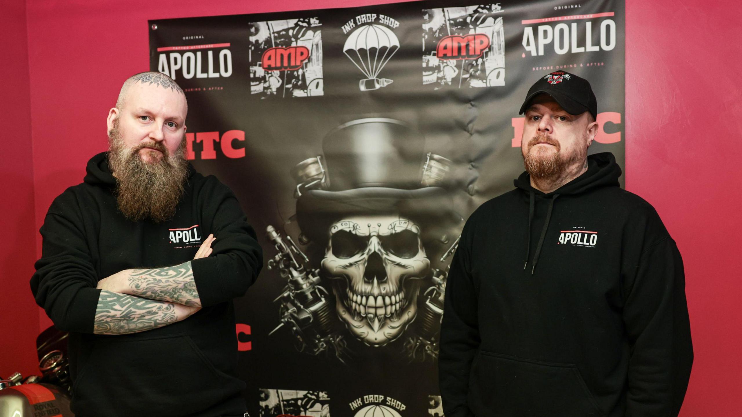 Stockport Tattoo Convention founders Chris Dodd (left) and Mike Gribben (right). Both men have beards and stand in front of a large black-and-white poster of a smiling skull wearing a top hat.