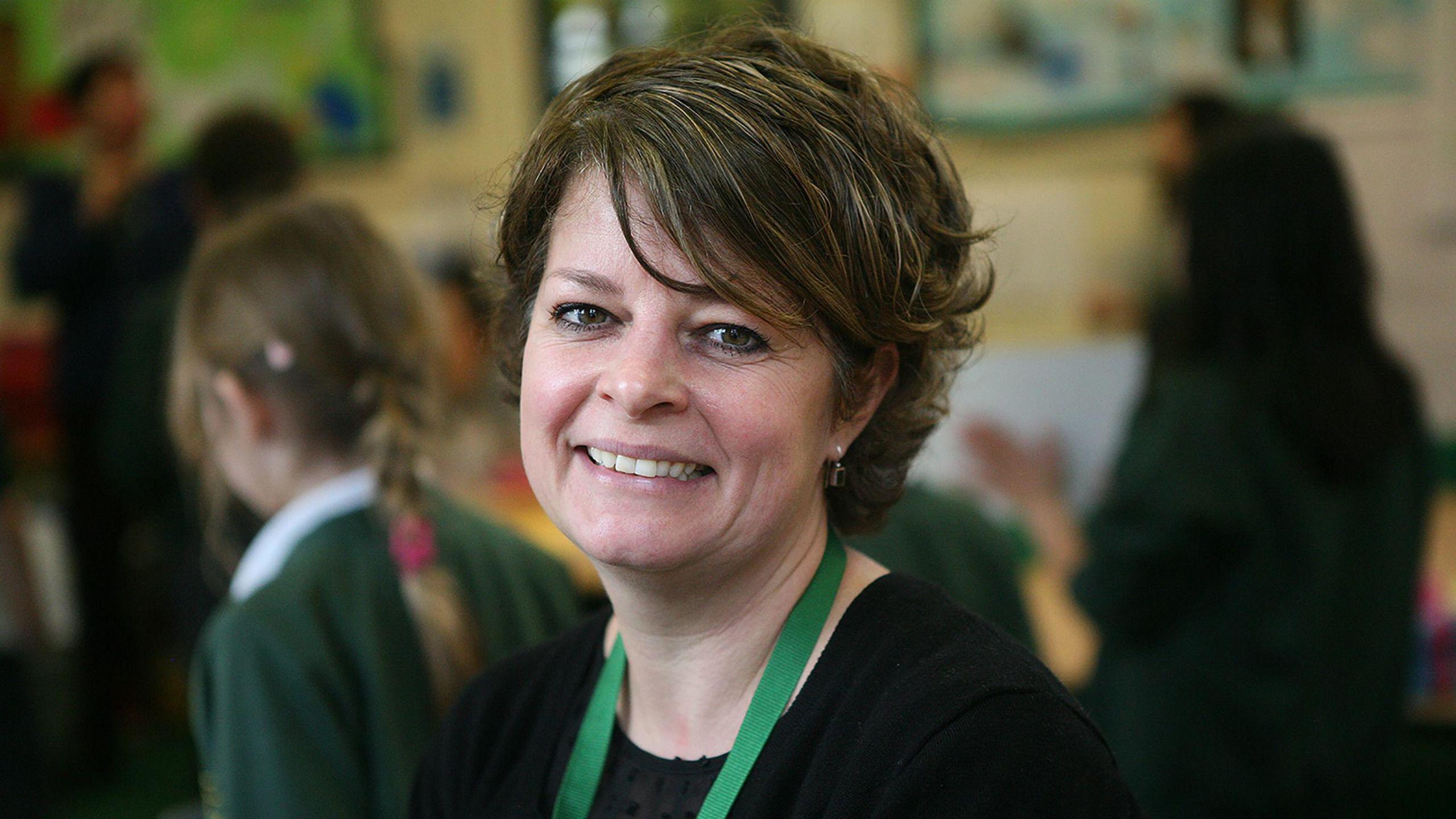 Ruth Perry. She has medium length brown hair, and is wearing a green lanyard around her neck. Out of focus in the background is a classroom full of children.