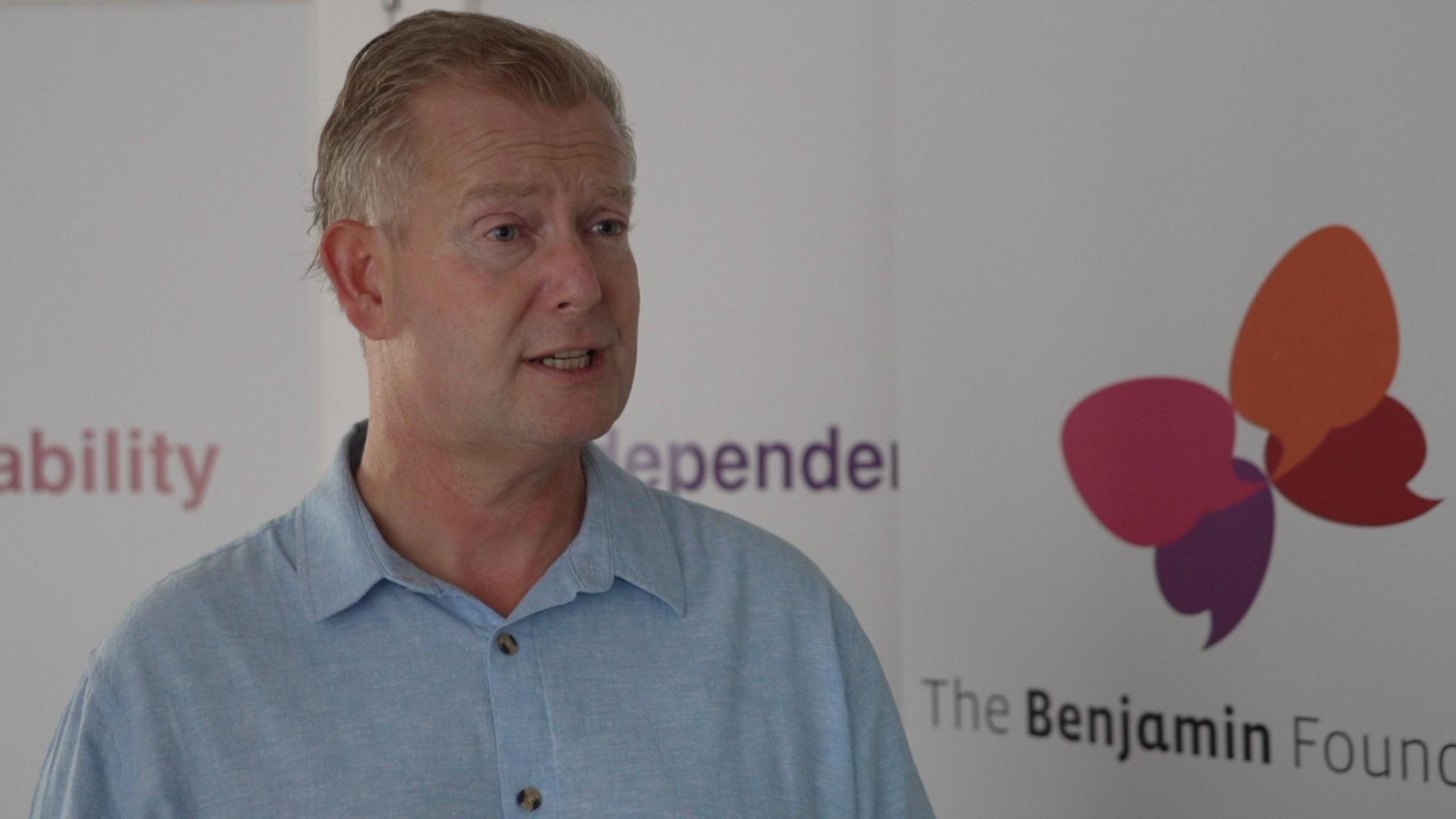 Matt Garrod, wearing a light blue polo shirt, looks off-camera, standing in front of a board that says "The Benjamin Foundation" and has a logo featuring a multi-coloured speech bubble logo.