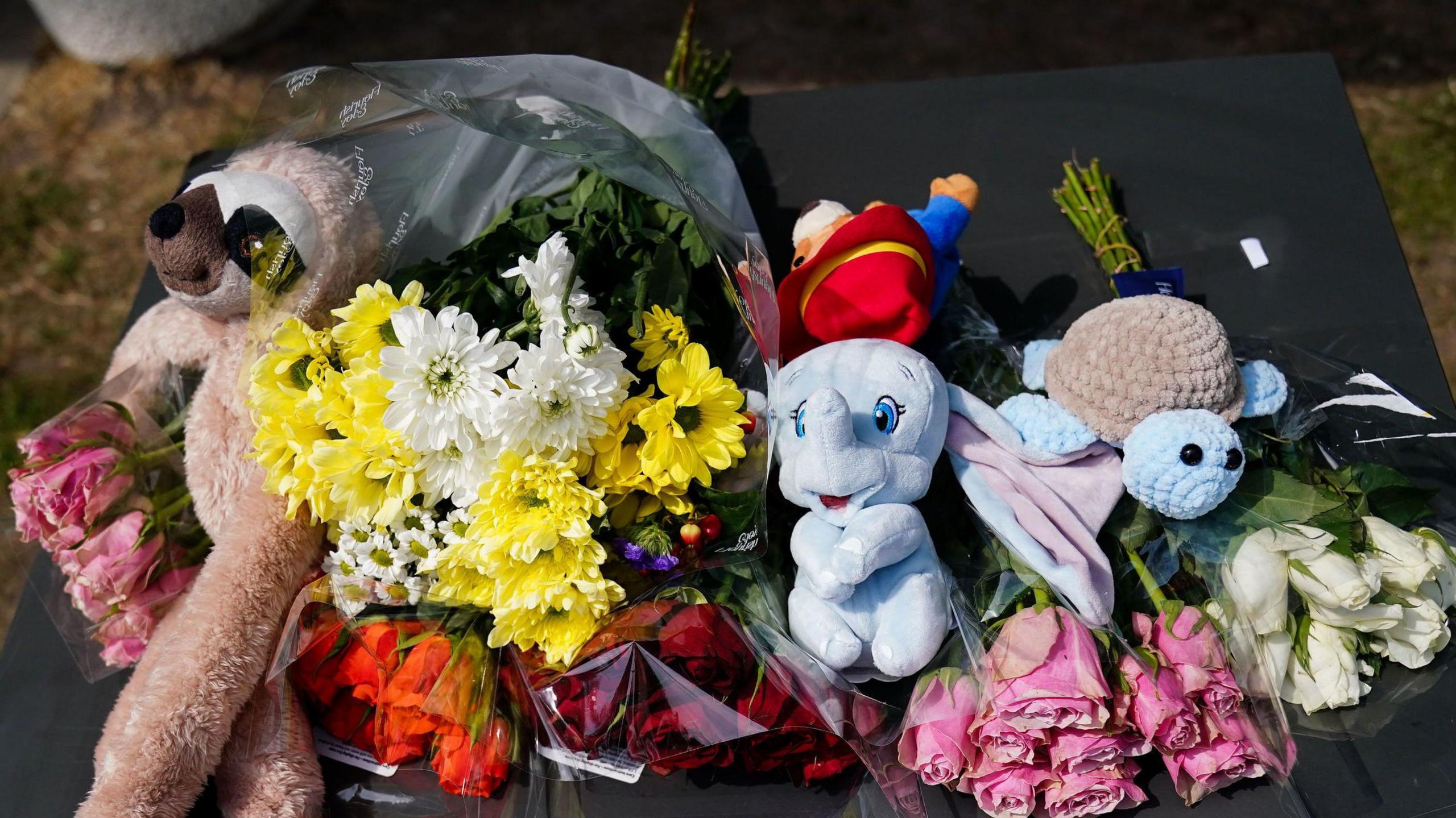 Flowers and cuddly toys arranged at the site. There is a toy elephant, turtle and monkey and several bunches of flowers.