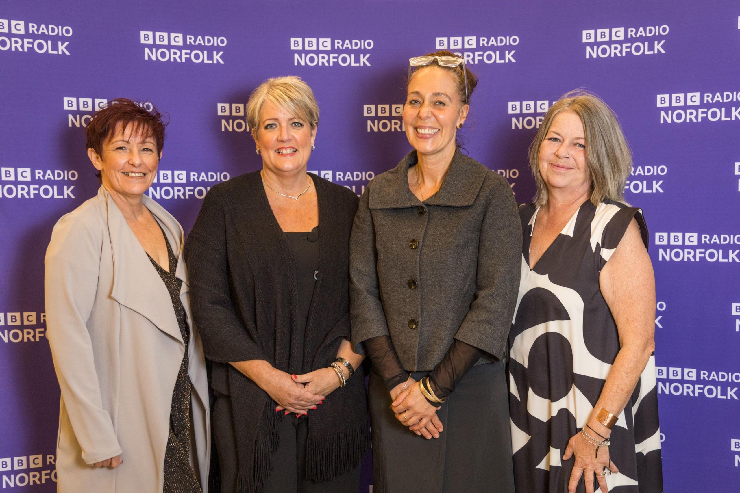Four women stand in front of a purple background. They are wearing mostly black dresses. They are smiling.