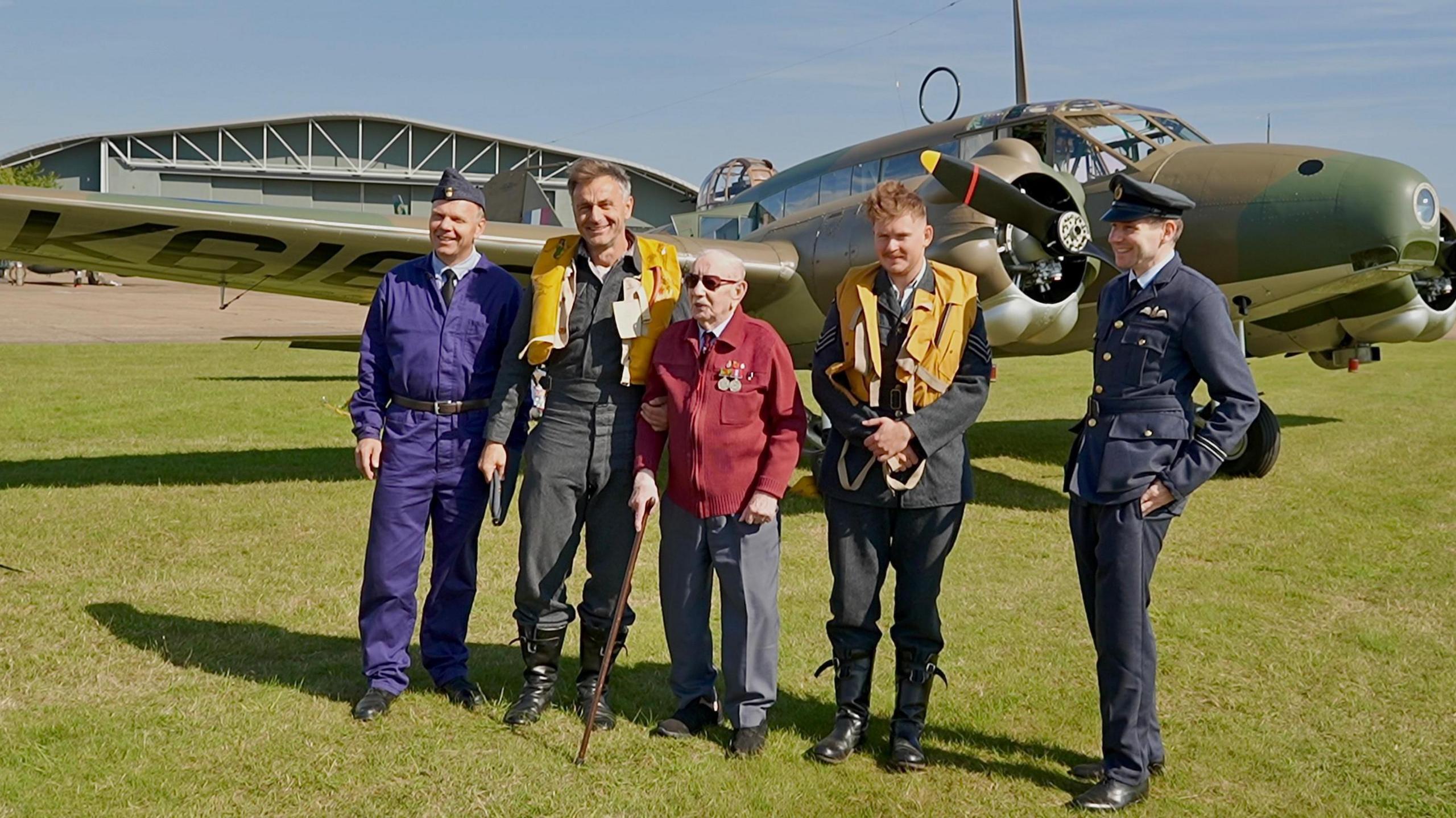 A group of people stood on the airfield in front of the Anson Mk.I with Mr Brown and Mr Santus in the centre. Mr Brown is standing using a stick and has medals pinned to his jumper. The four men he is standing with are all wearing vintage looking RAF uniforms. Two of them - including Mr Santus - have what looks to be yellow life jackets over the top of their uniform.