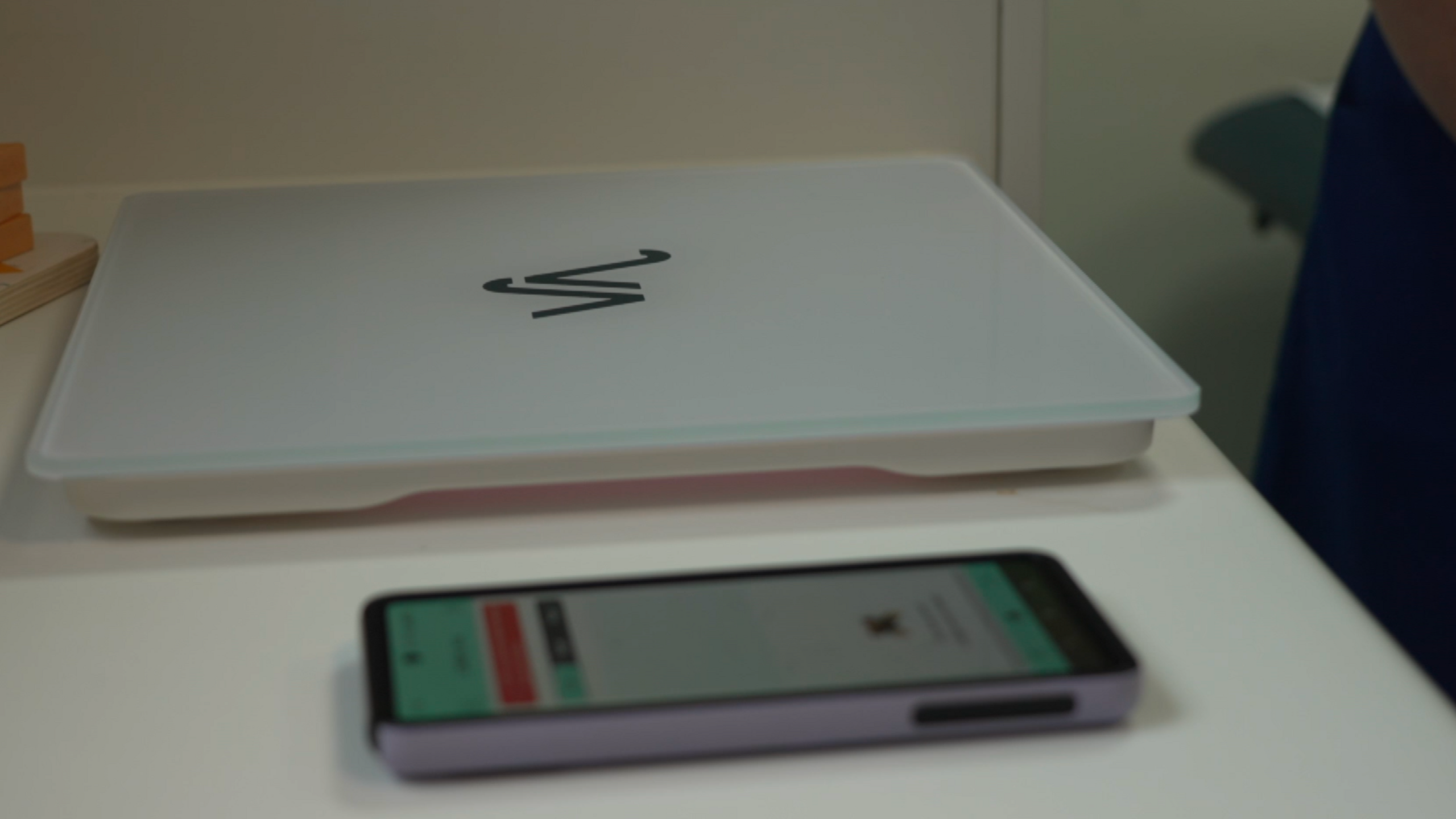 Smart scales - a flat surface for weighing children - sit on a table next to a mobile phone featuring the app which the scales link to