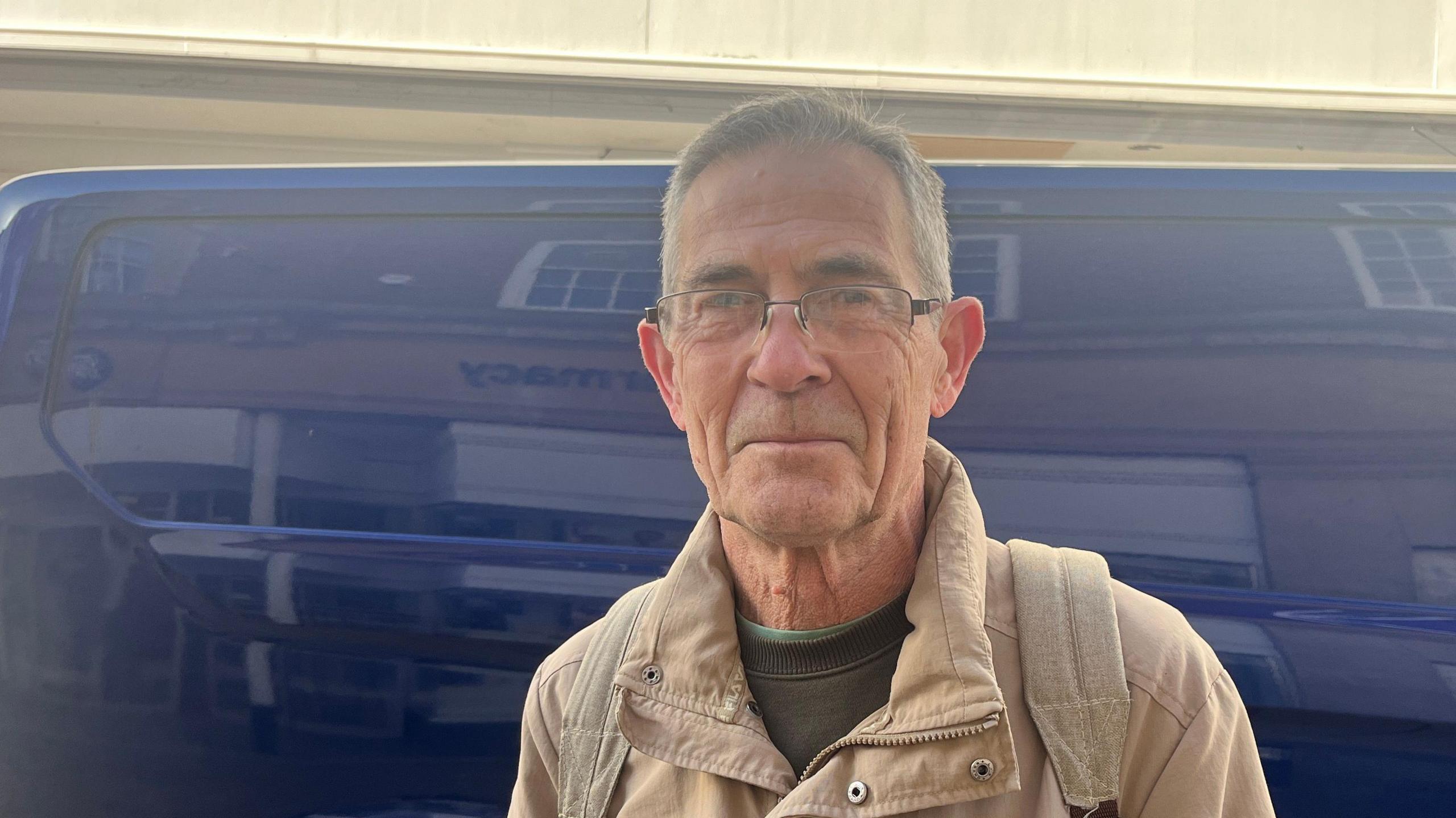 Paul Jackson is wearing a cream coat over a dark green jumper. He is wearing glasses. The reflection of a passing bus is behind him.