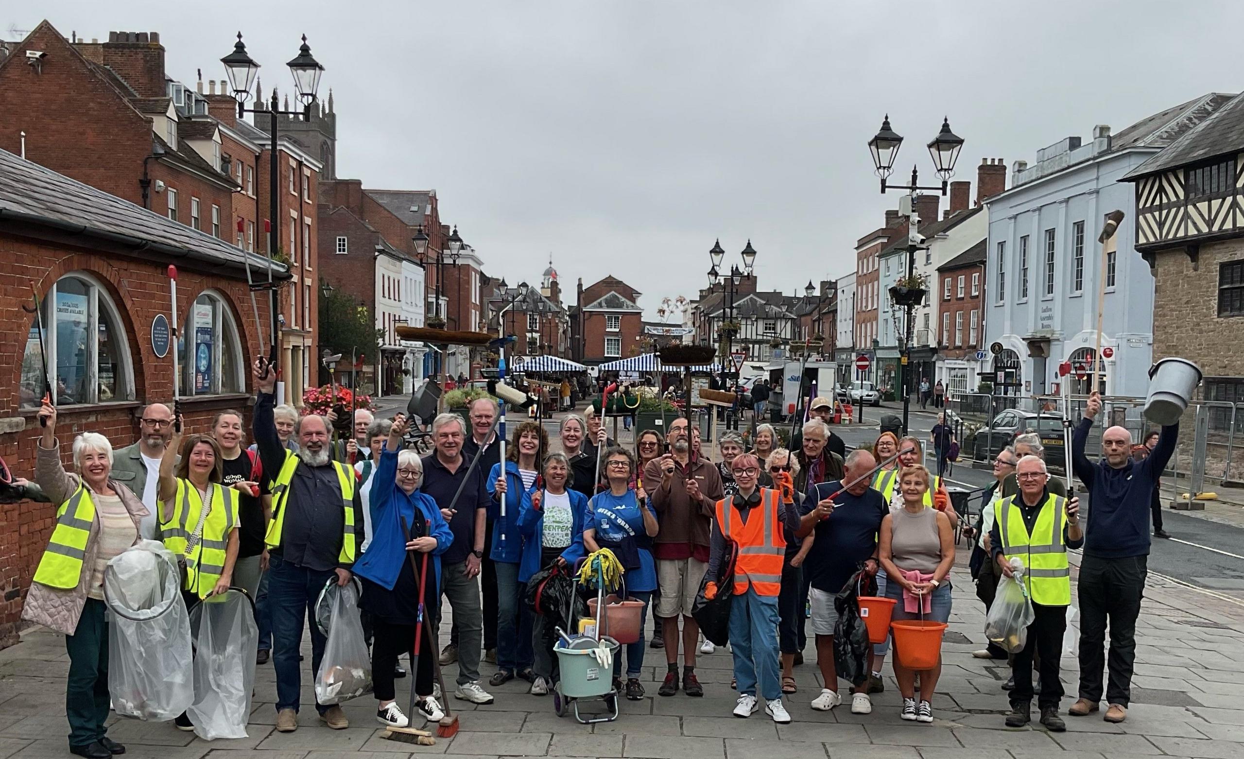 About 30 people, some in high-vis jackets, pose for the camera in the foreground. Many are carrying cleaning equipment. Buildings are in the background.