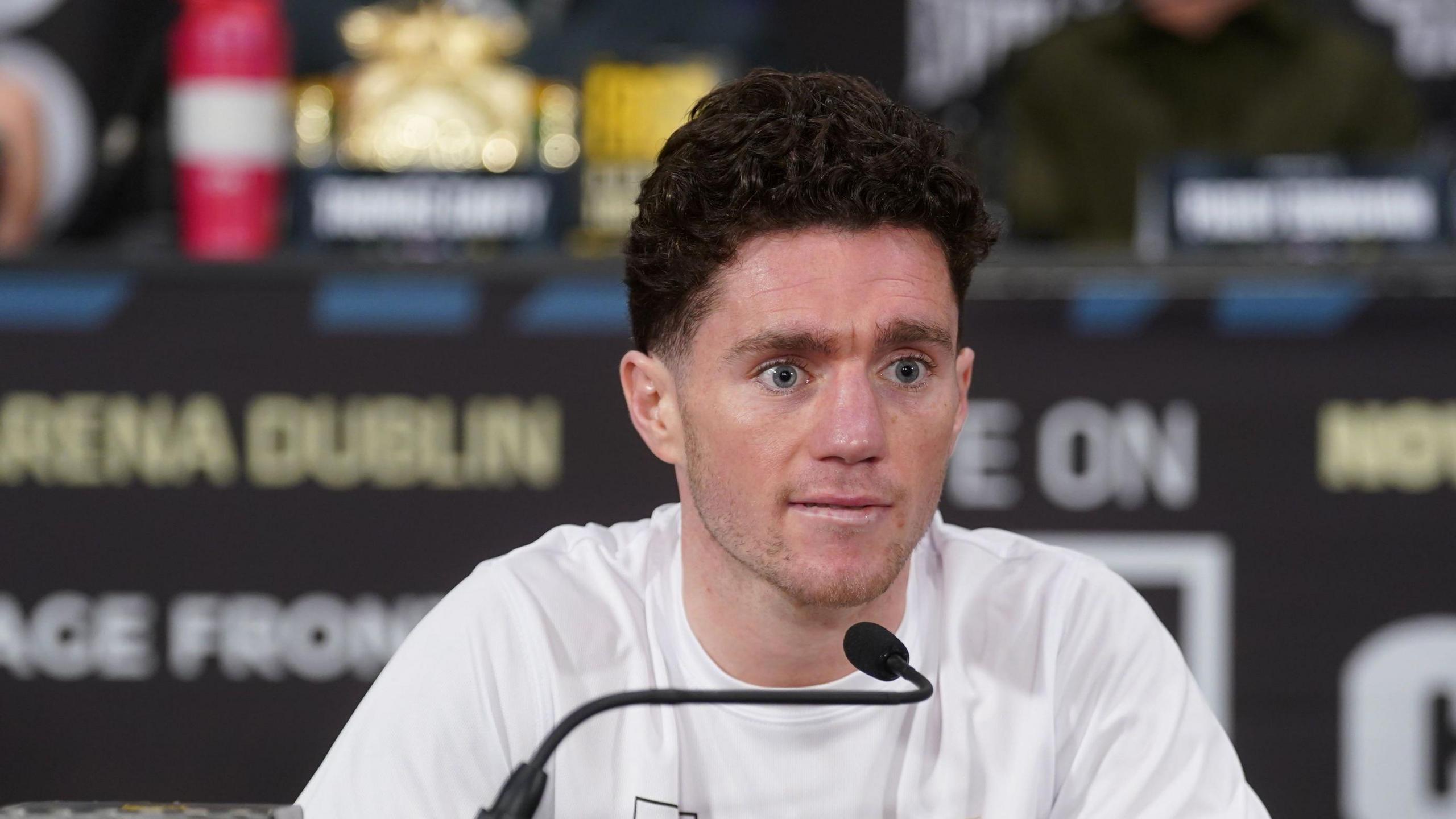 The late John Cooney pictured at a press conference.   He has short, dark, curly hair and blue eyes and is wearing a white t-shirt.   He is sitting in front of a microphone. 