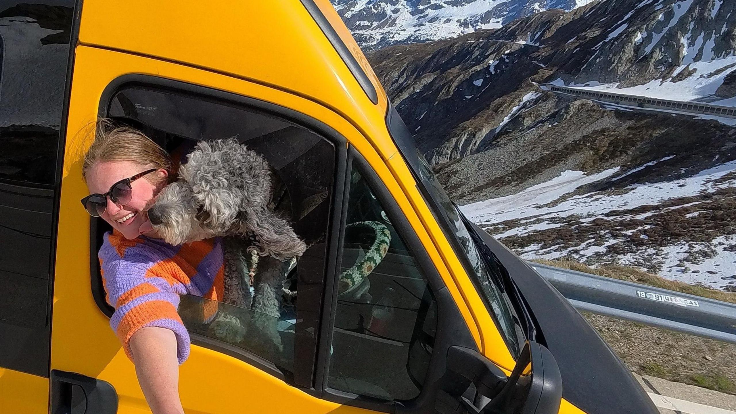 A selfie taken by Ella Dungey, showing her sitting in the driver's seat of her yellow and black van with her dog on her lap. She is smiling and wearing sunglasses and a colourful jumper. In the background are snowy mountains and a road winding through them.