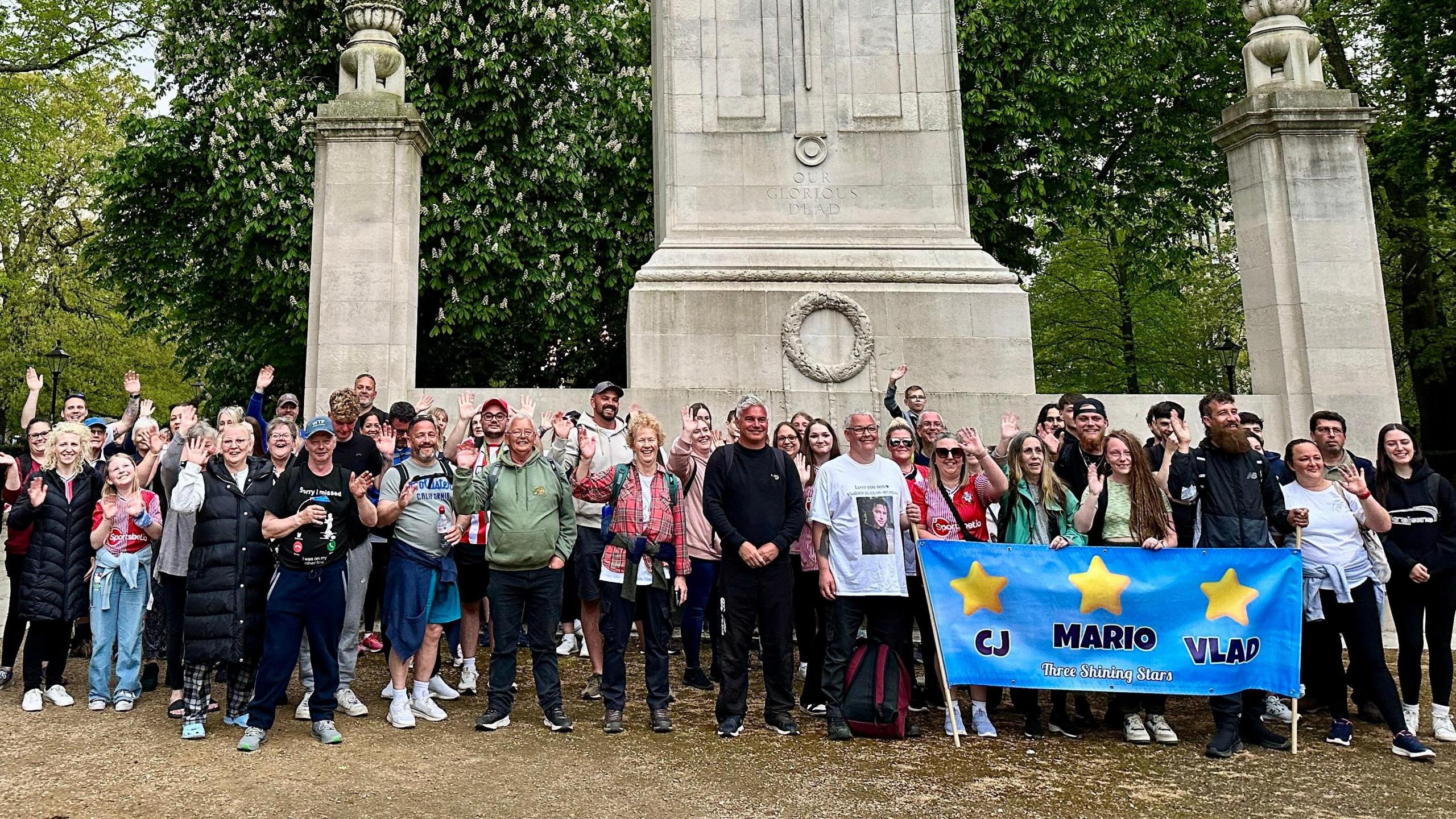 About 80 walkers are pictured standing in front of Southampton Cenotaph in the city centre. They are wearing comfortable hiking clothes and smiling and waving to the camera. A blue banner inscribed with the names CJ, Mario and Vlad is being held by their families.