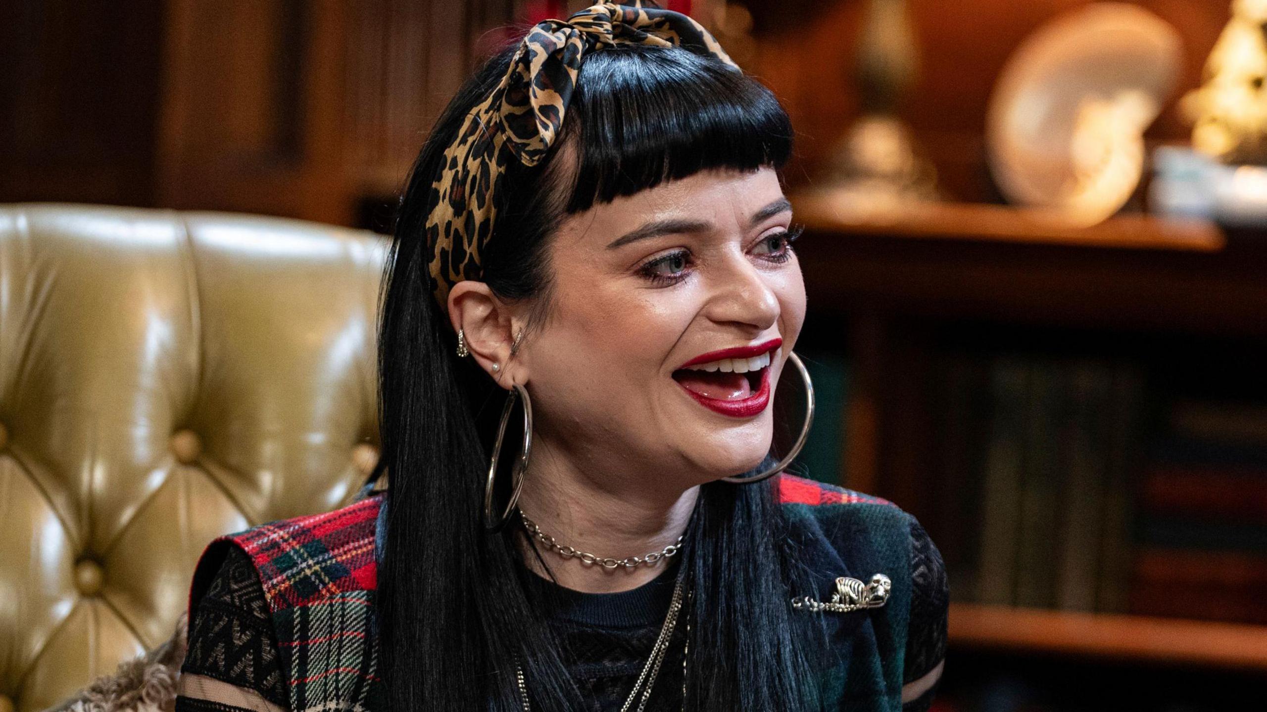 Ruth Codd is smiling on camera on The Celebrity Traitors. She has a leopard-print bow in her hair and tartan patterned clothing across her shoulders. She is wearing lots of jewellery including large looped earrings. She has bright red lipstick and jet black hair with a fringe hanging sharply on her forehead.