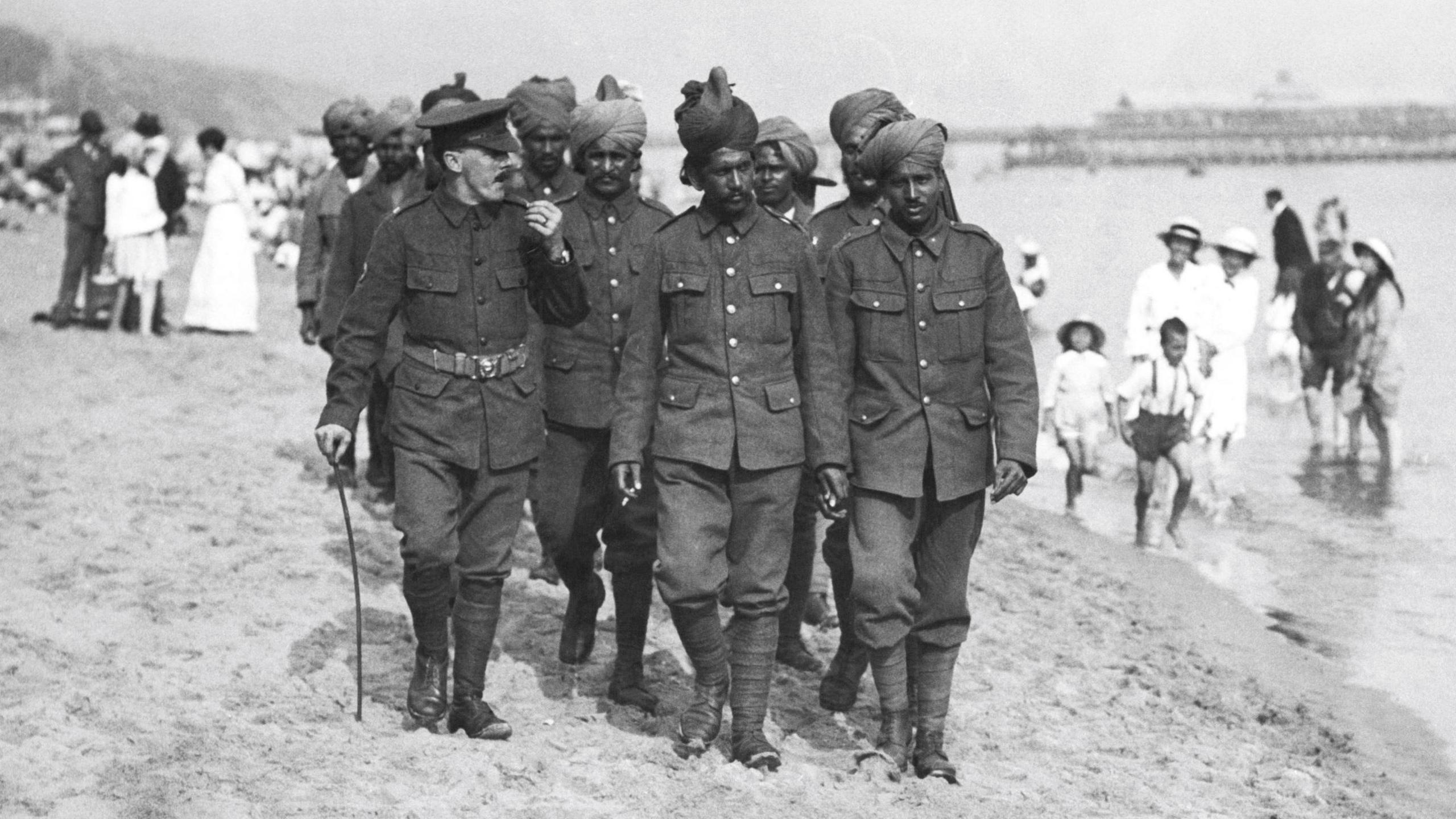 In a black and white photo from 1915, a dozen or so Indian soldiers walk on Bournemouth beach, accompanied by a British officer. The pier is in the background and people are paddling in the sea.