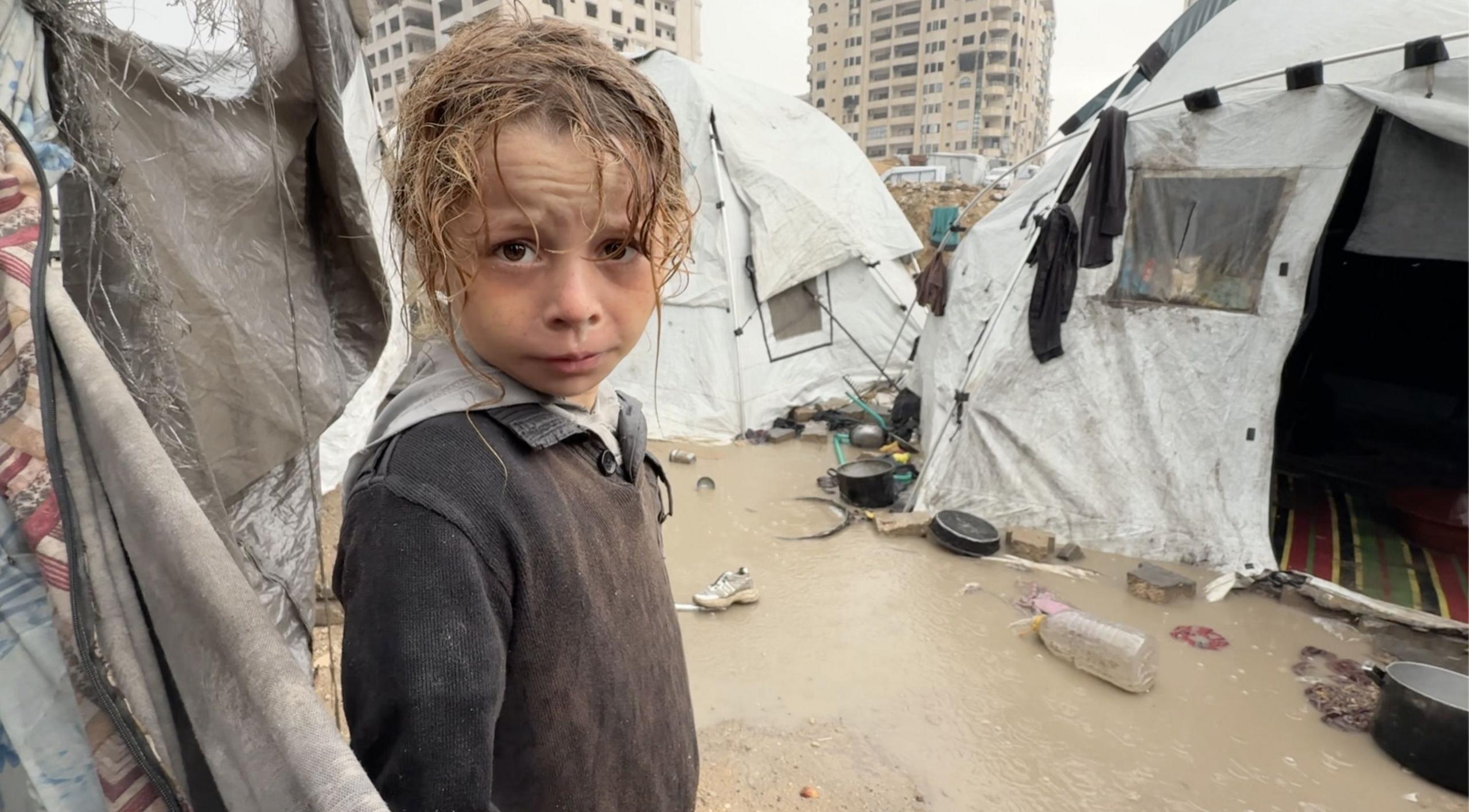 A Palestinian child, wet from rain, looks into the camera, surrounded by soaked tents and wet and muddy ground.