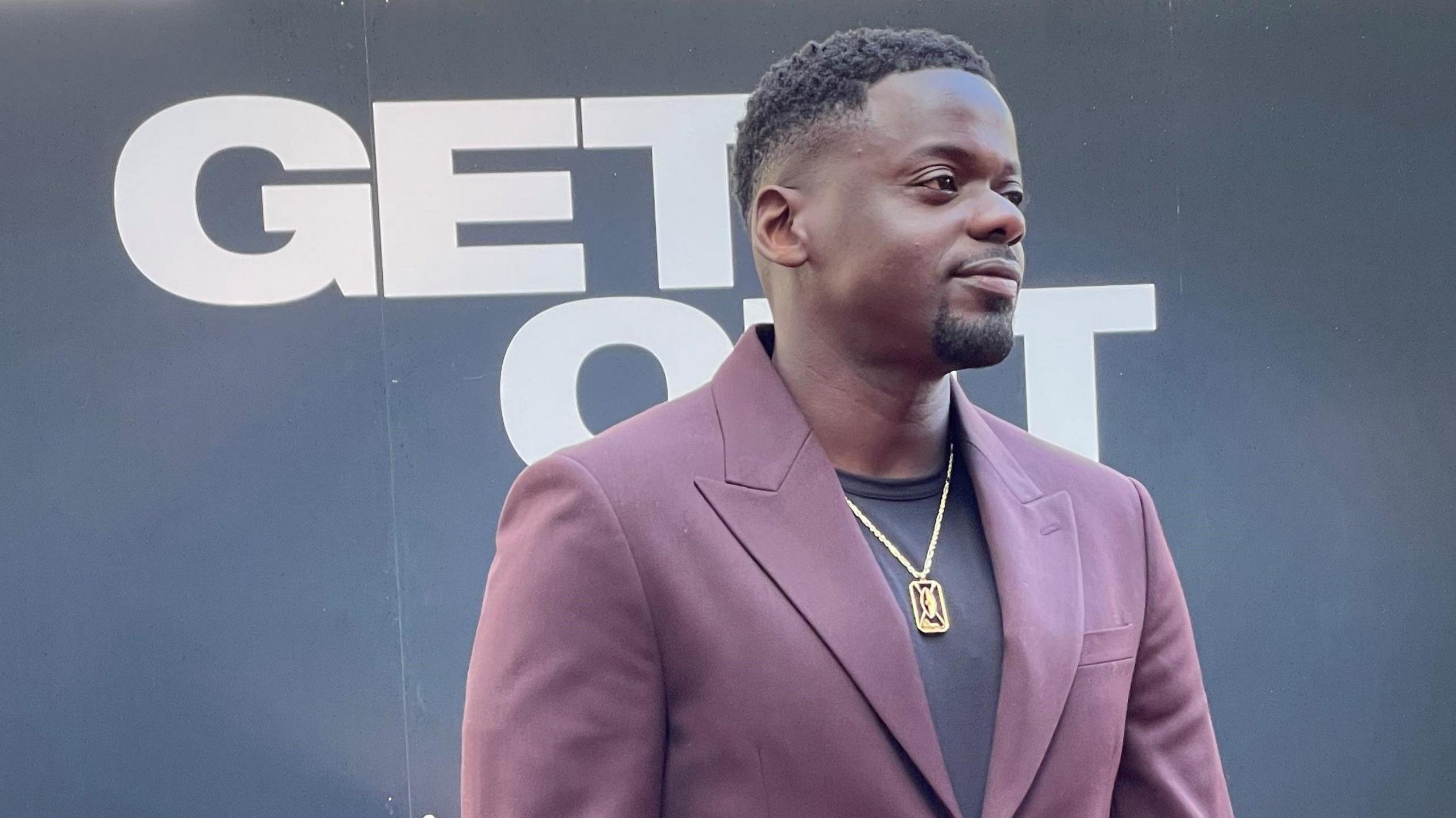 Actor Daniel Kaluuya wearing a mauve suit and dark blue t-shirt and a decorative necklace stands in front of a Get Out sign. 