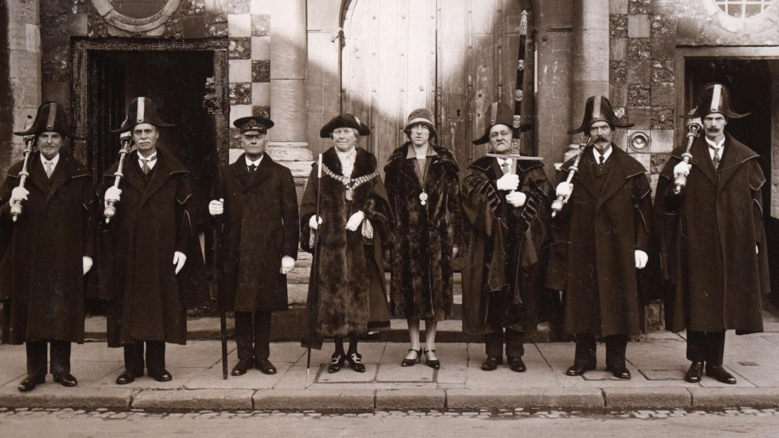 Archive image of Florence Ada Coxon from the 1920s dressed in her ceremonial robes and hat. To her right is her daughter who was her mayoral consort. They are in a line of men dressed in ceremonial gear. They are all dressed in long dark coats and big hats. 