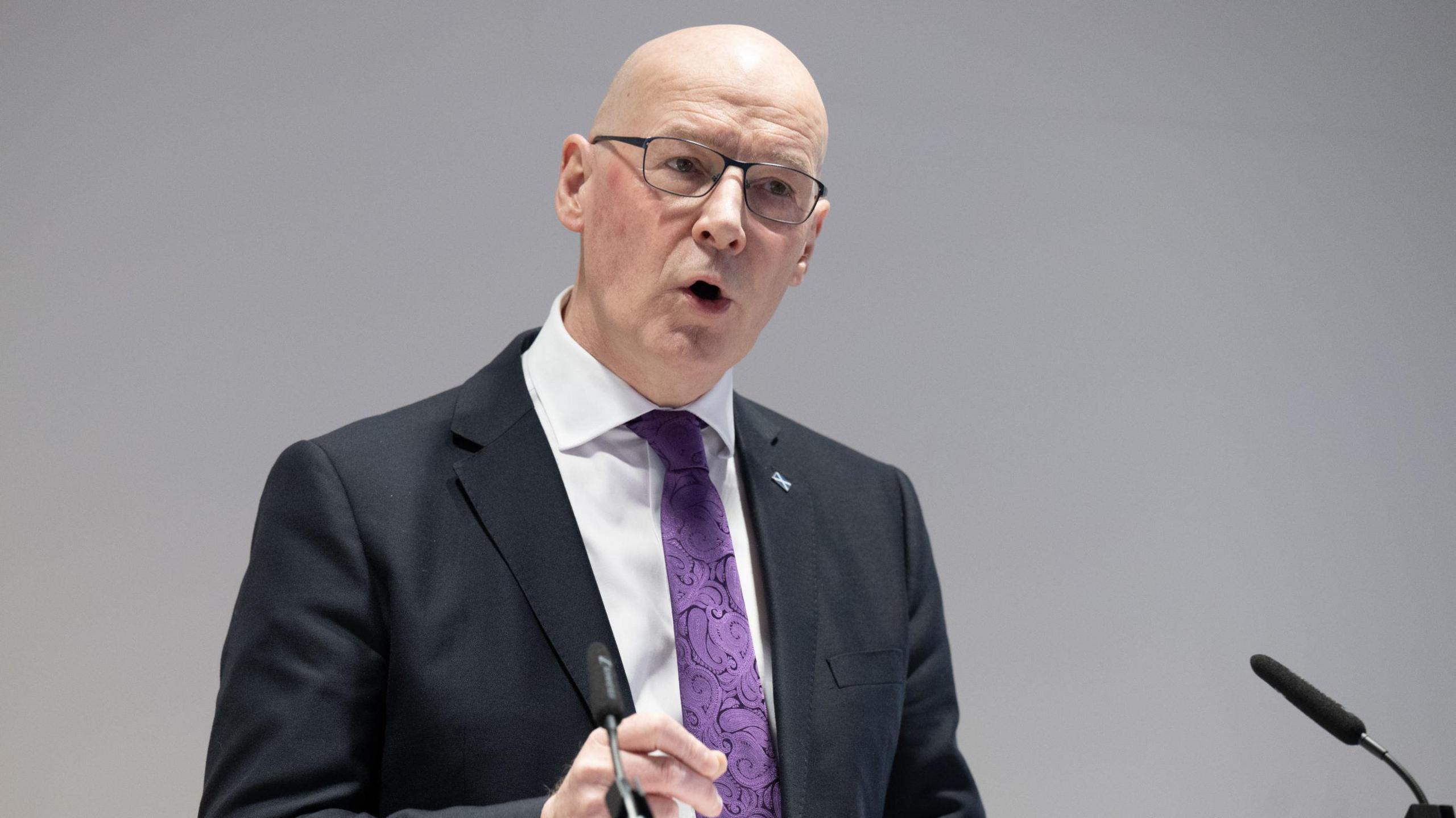 John Swinney speaking onstage while giving a speech. He has a black jacket, white shirt and purple tie on.