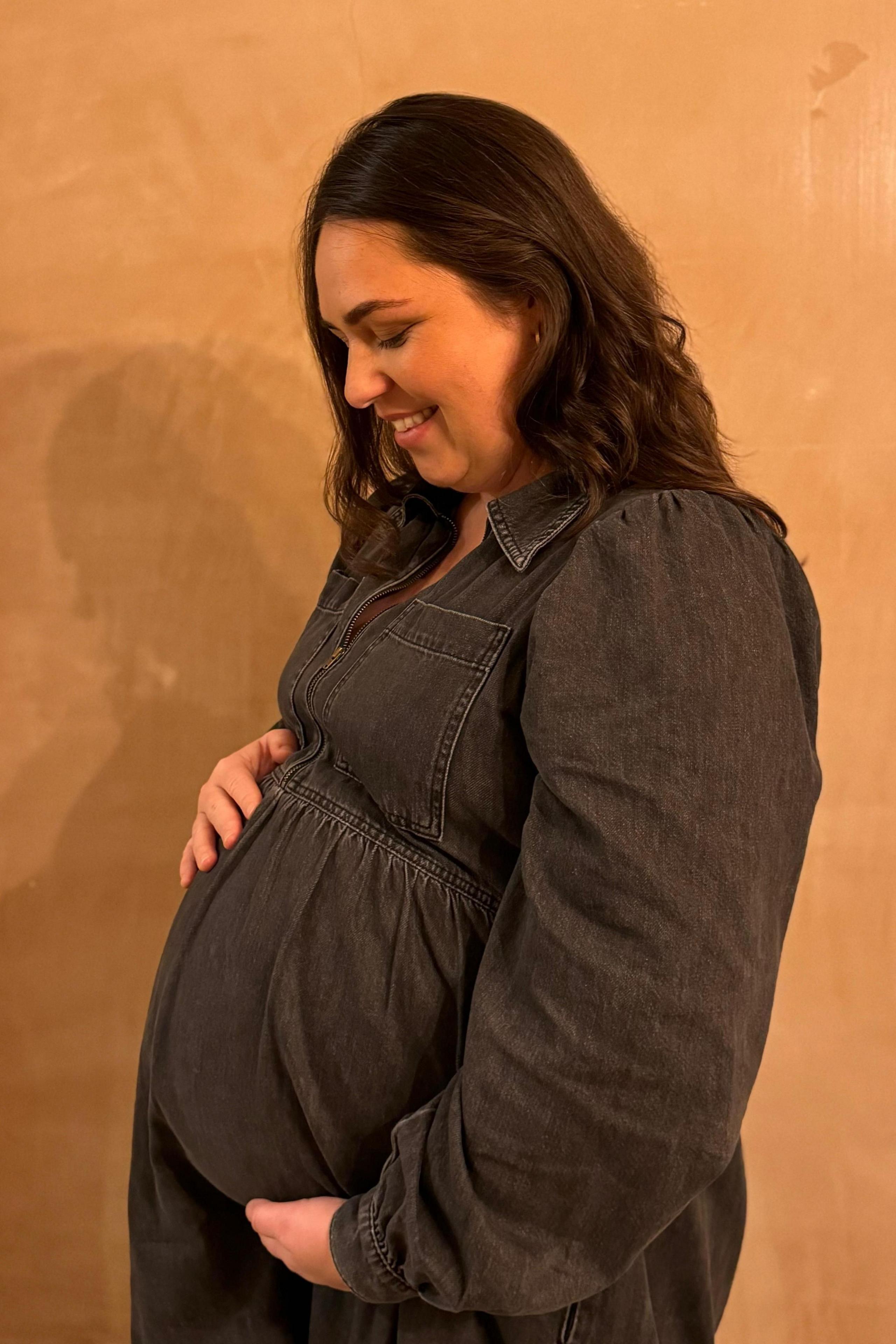 Amy, wearing a black denim tunic, is pictured side-on in front of a tan-coloured wall. She is holding her baby bump between both hands and is looking down and smiling.