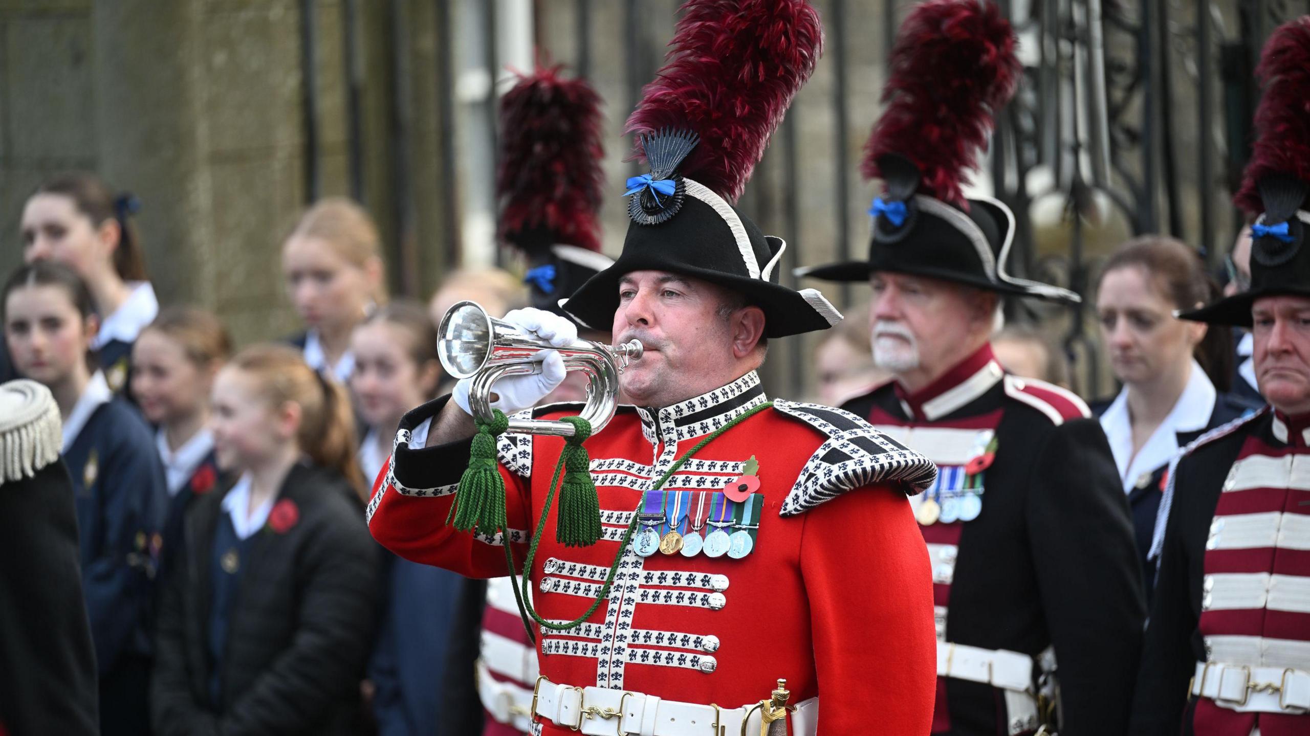 A man, who's wearing a red army jacket with medals and a feather sticking out of his hat, is playing a trumpet. He's standing with a crowd about him.