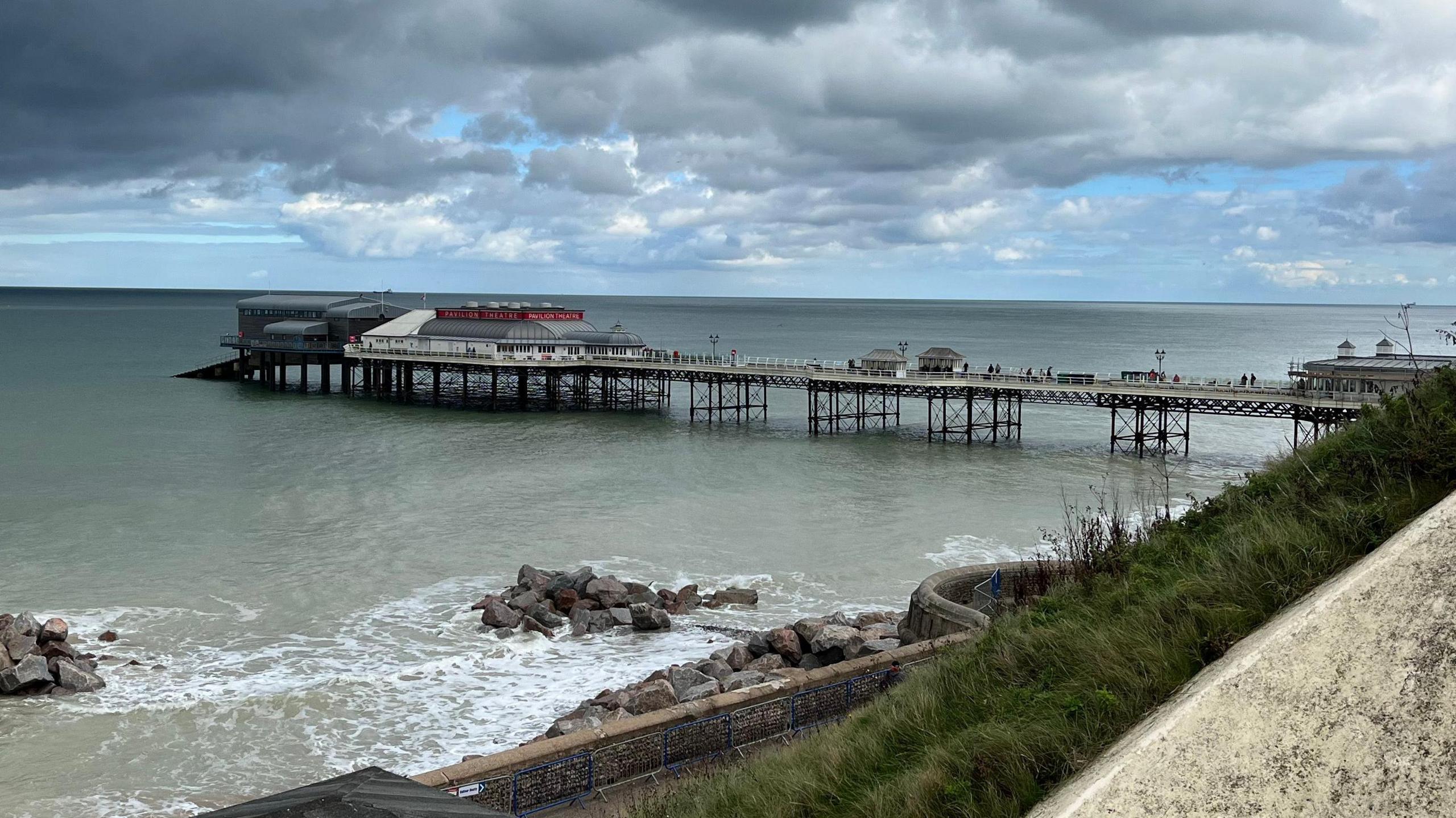 A view of a promenade pier with a building at the end of the structure. The pier juts out into the sea with a few rocks gathered to the structure's left. 