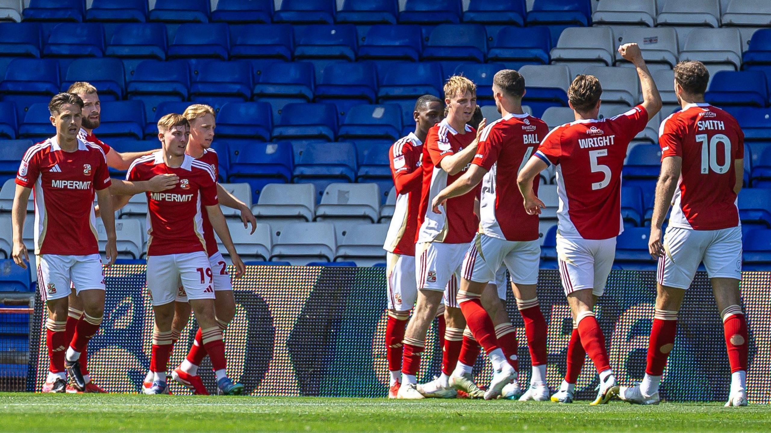 Players from Swindon Town FC, in the club's traditional kit of white shorts and red shirts, group together as they celebrate a goal at Oldham Athletic. In the background is a grandstand with blue and white seats, most of which are empty