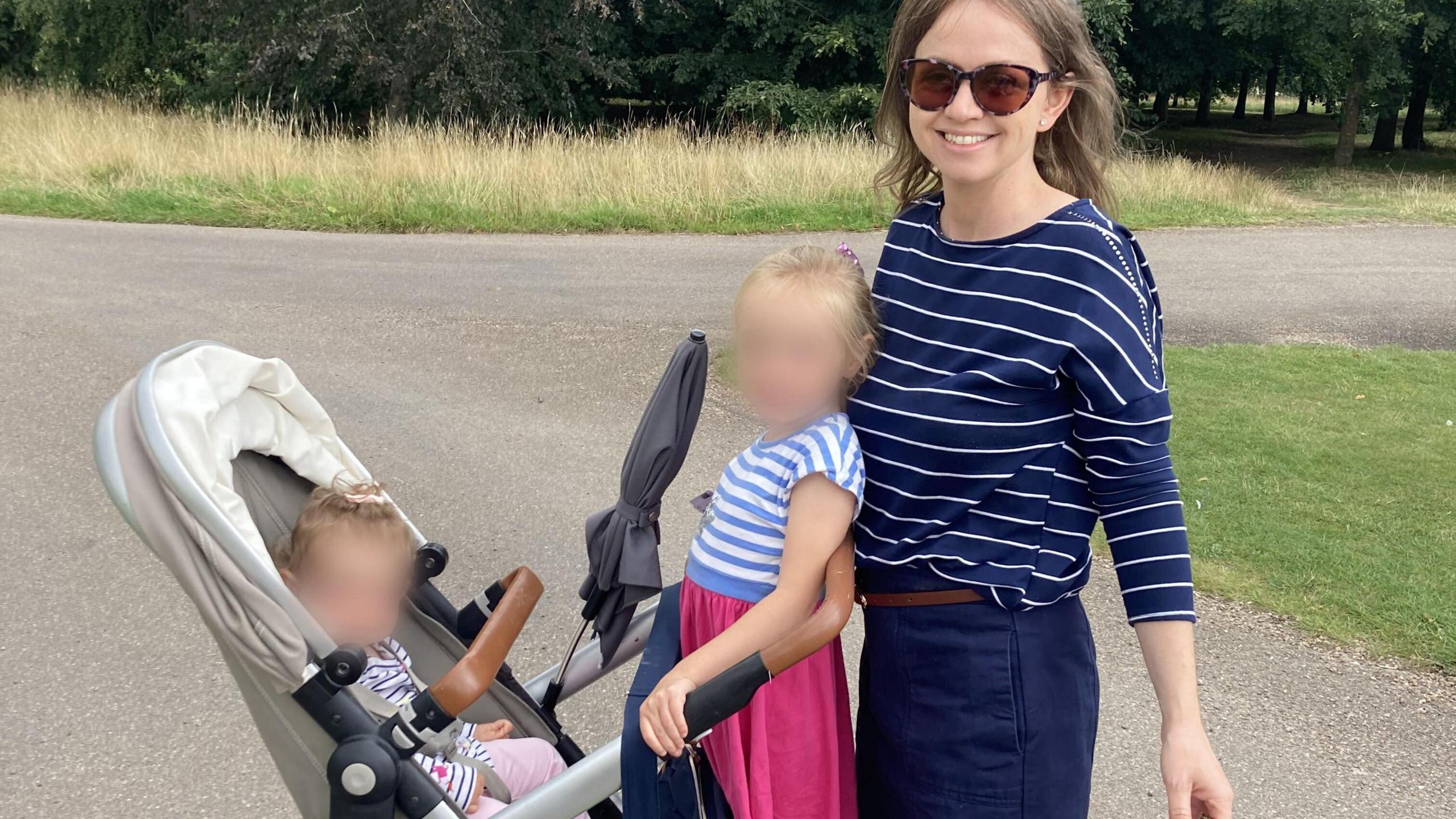 A woman in a park with two children. She has dark hair and is wearing sunglasses, a blue and white striped top, and blue trousers. The woman is standing in front of a buggy - there is a child standing on the back of the buggy who has blonde hair and is wearing a blue and white striped top and a pink skirt. Her face is blurred out. Sitting in the buggy is a toddler whose face is also blurred. Behind them is trees and grass.