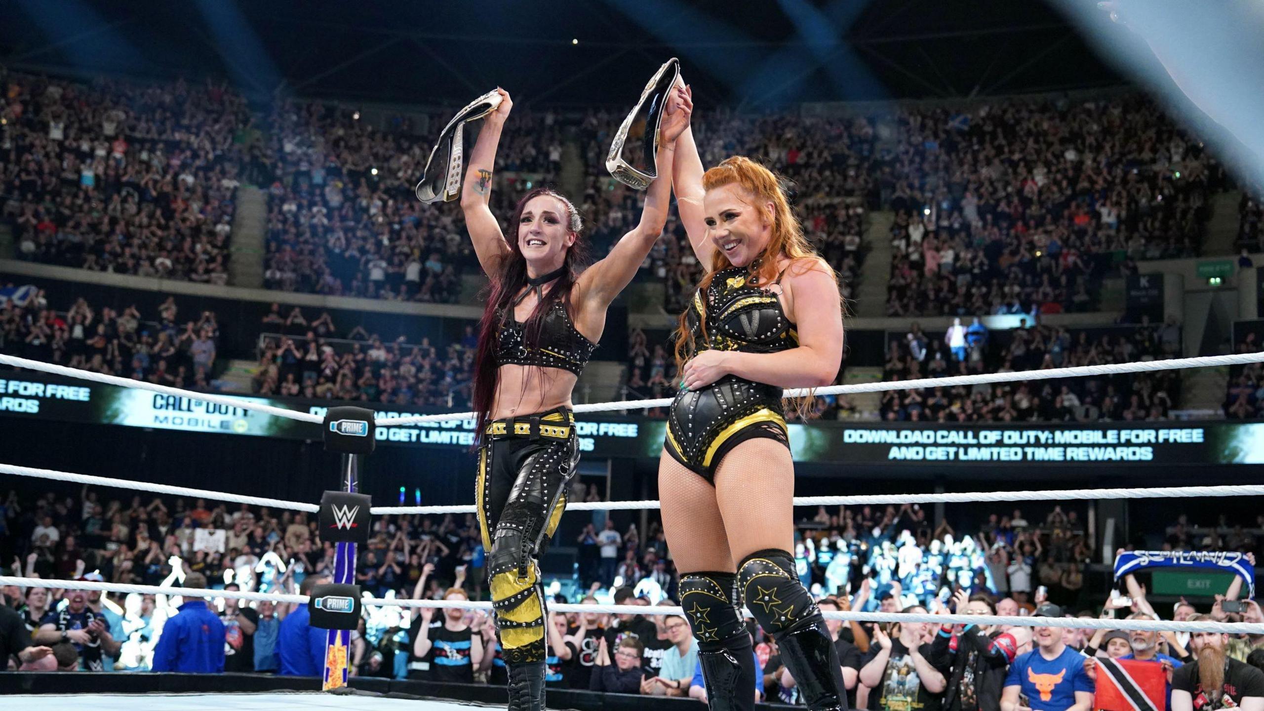 Alba Fyre and Isla Dawn celebrate winning the tag team titles at Clash at the Castle.  Both women are grinning and standing in the centre of the ring, holding their titles high. They are wearing black and yellow ring gear. Fans in the arena are celebrating behind them.