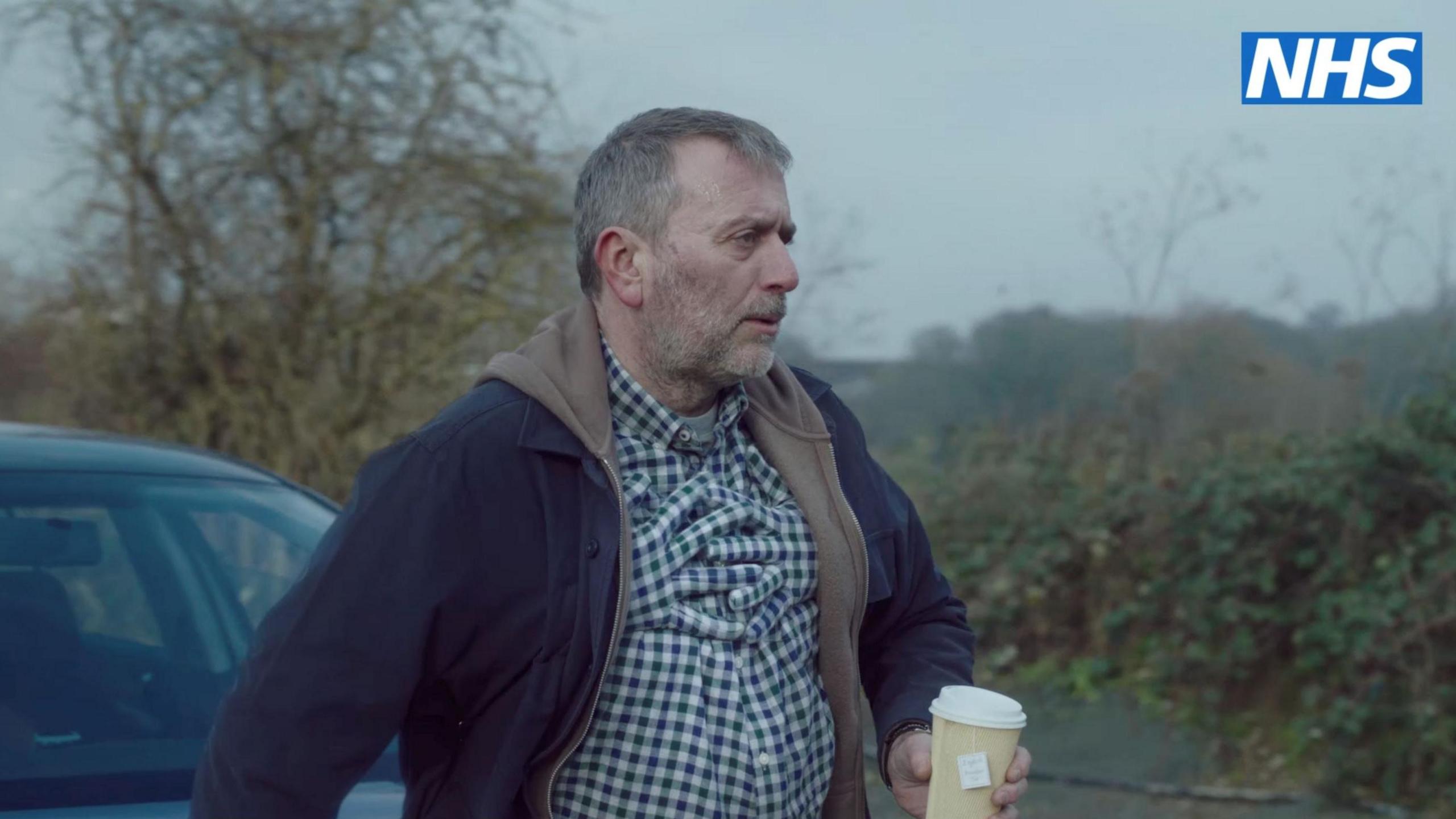 Robert in the NHS video, which shows arms around him, to stimulate how the tightening of a chest can feel like, he is wearing a blue and green checked shirt, he is holding a cup of coffee and is outside by a car. 