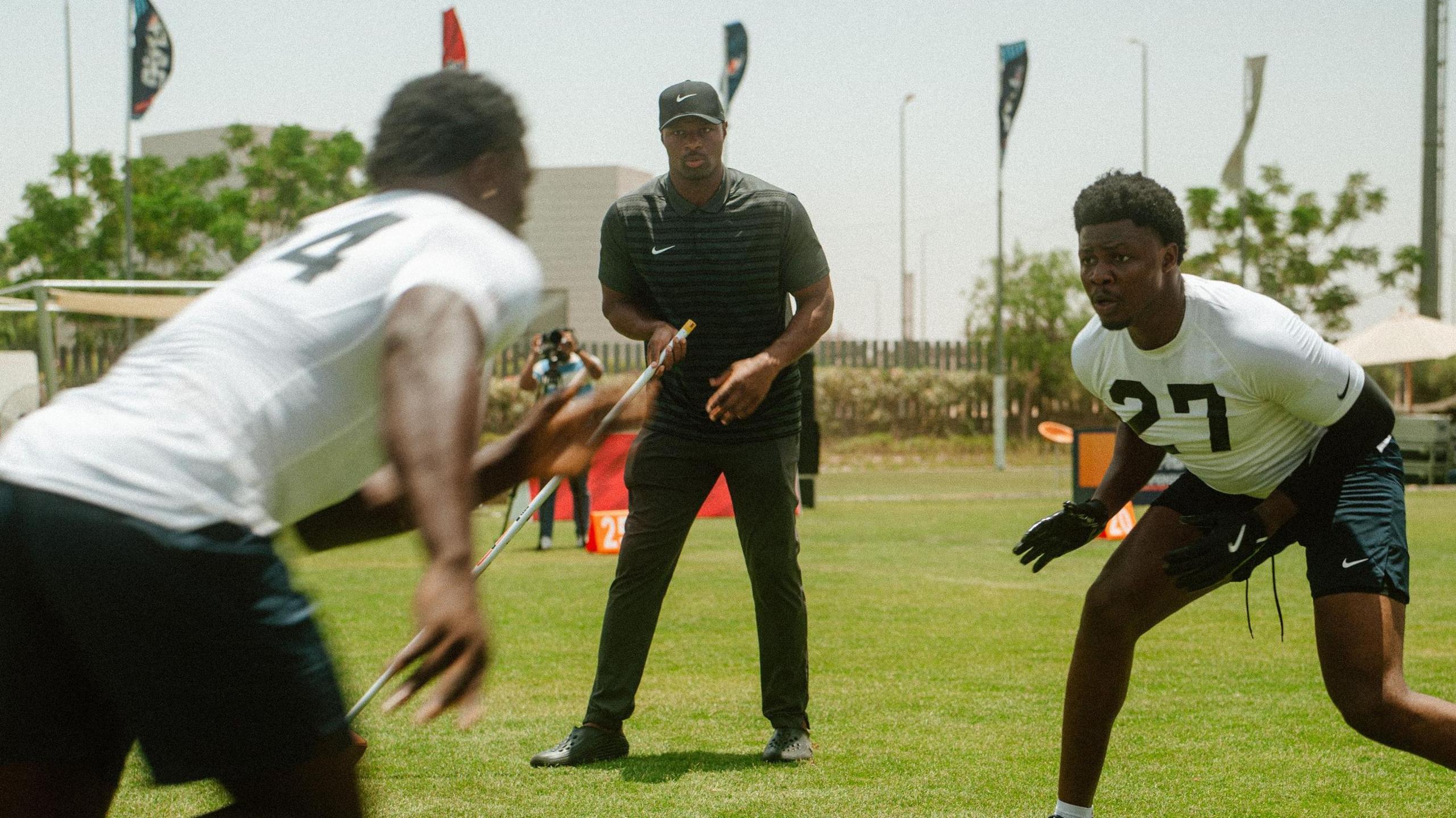 Osi Umenyiora, wearing a black baseball cap, a black t-shirt and grey trousers, stands on an American Football training field holding a silver pole in his right hand as he watches two players in action. One player is running from the left of shot towards a defender on the right of shot, who is crouched down slightly and about to attempt to intercept him. Both players wear white t-shirts and black shorts